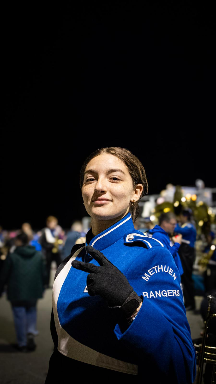 A young woman in a blue marching band uniform making a peace sign with her right hand, at a nighttime band event with group members blurred in the background.