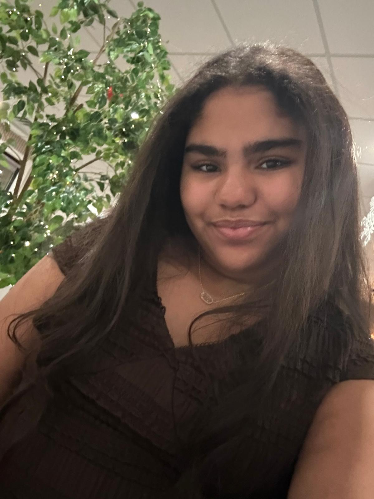 A young woman with long dark hair wearing a dark brown top and a delicate necklace, smiling at the camera. In the background, there is a tall houseplant with green leaves and small lights, and a ceiling with white tiles.