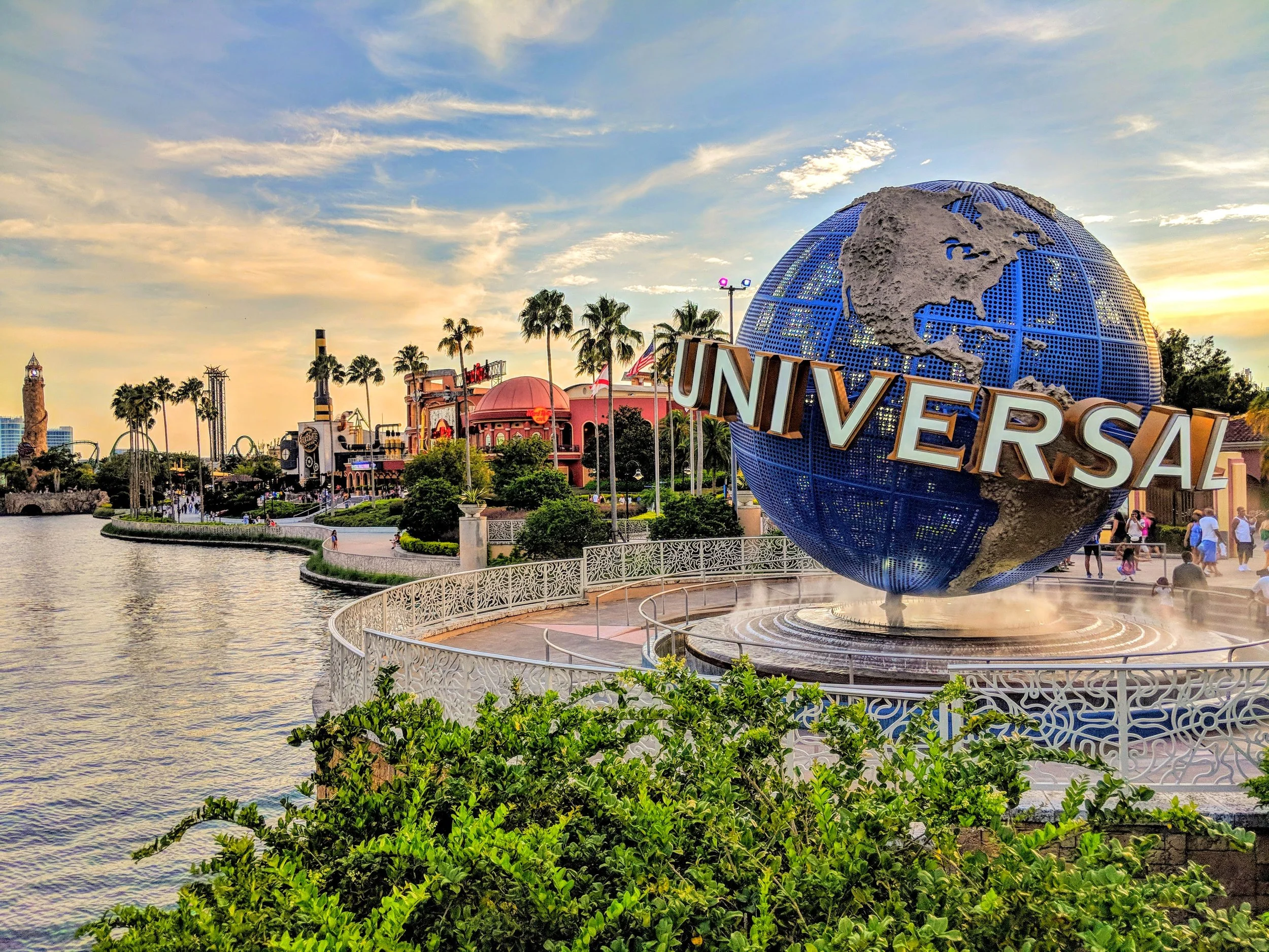The Universal Studios globe fountain at sunset with colorful sky, palm trees, and amusement park buildings in the background.
