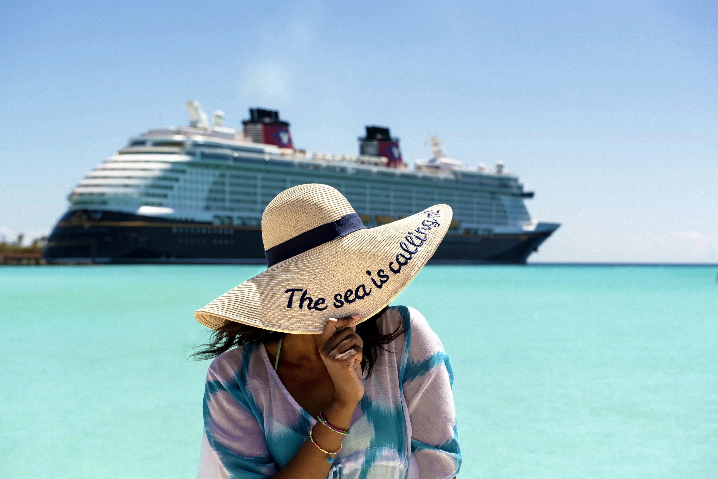 Woman wearing a large sunhat that says 'The sea is calling' on a tropical beach with a Disney Cruise Line ship in the background.