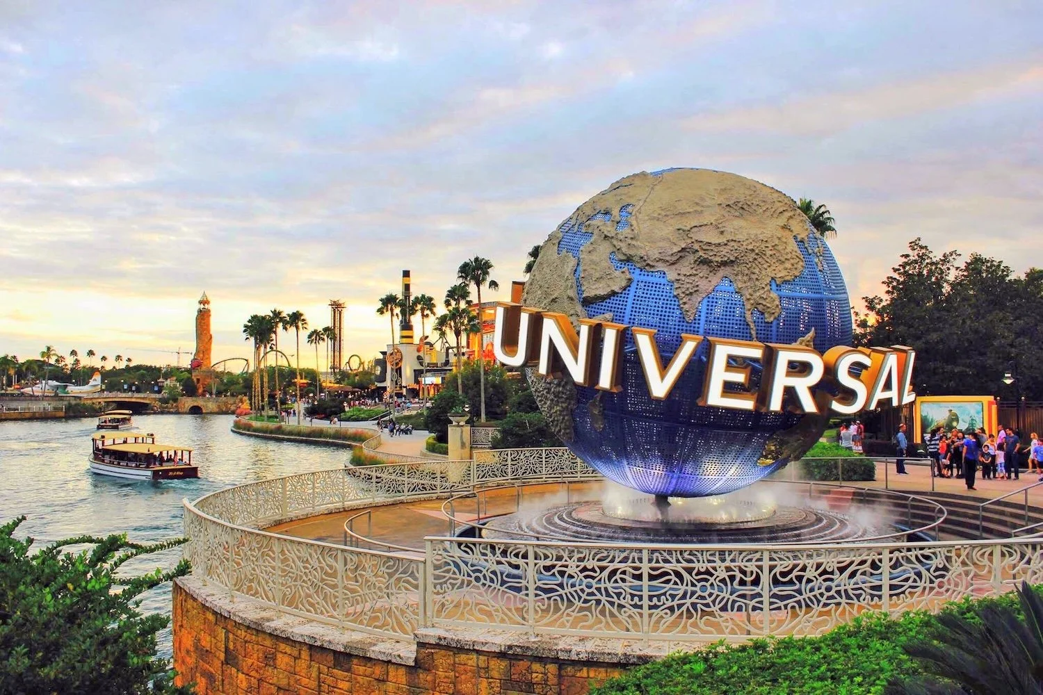 The iconic Universal globe at Universal Studios theme park with a water fountain at its base, surrounded by visitors and tropical trees, during sunset.