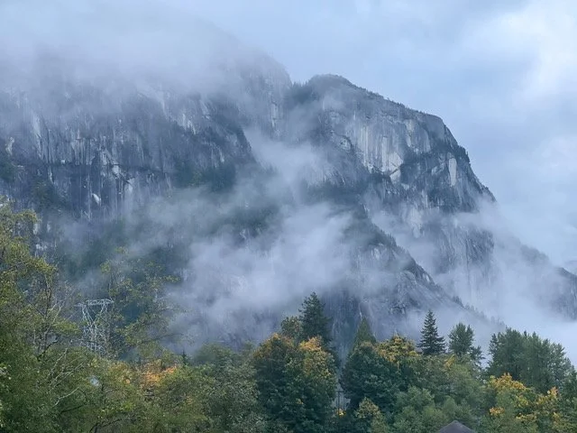 Mountain with rocky cliffs partially obscured by fog and surrounded by green trees.
