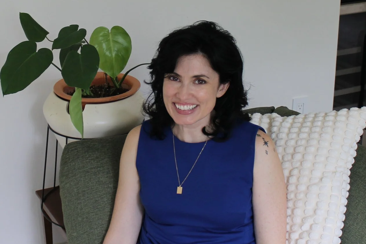 A woman with black hair, wearing a sleeveless blue dress and gold jewelry, sitting on a green sofa with a textured white pillow, smiling at the camera in a living room with a potted plant and kitchen cabinets in the background.