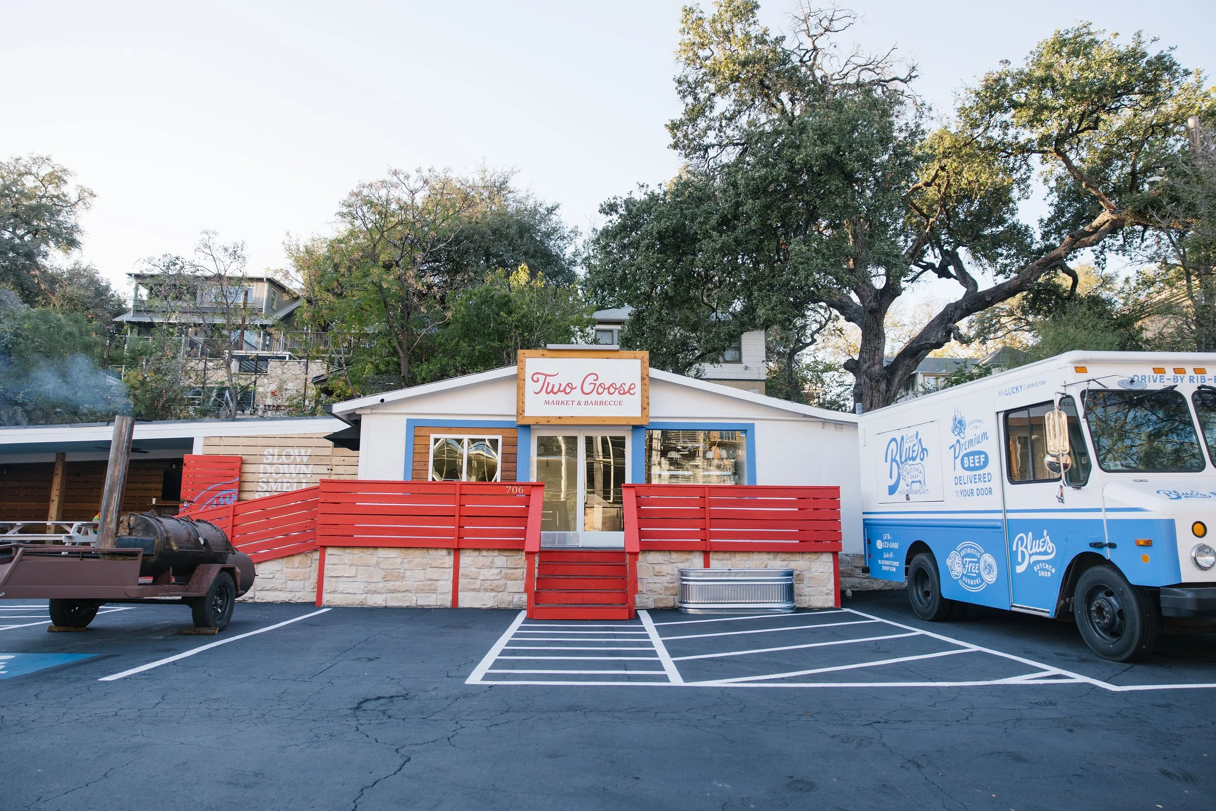 Small restaurant with a sign reading 'Two Goose Market & Barbecue,' red wooden stars at the entrance, parked food trucks including one with blue and white branding, and trees in the background.