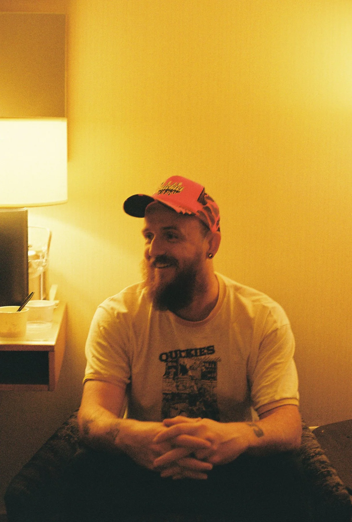 A man with a beard and earrings wearing a red cap and a white t-shirt, sitting at a table in a warmly lit room, smiling.