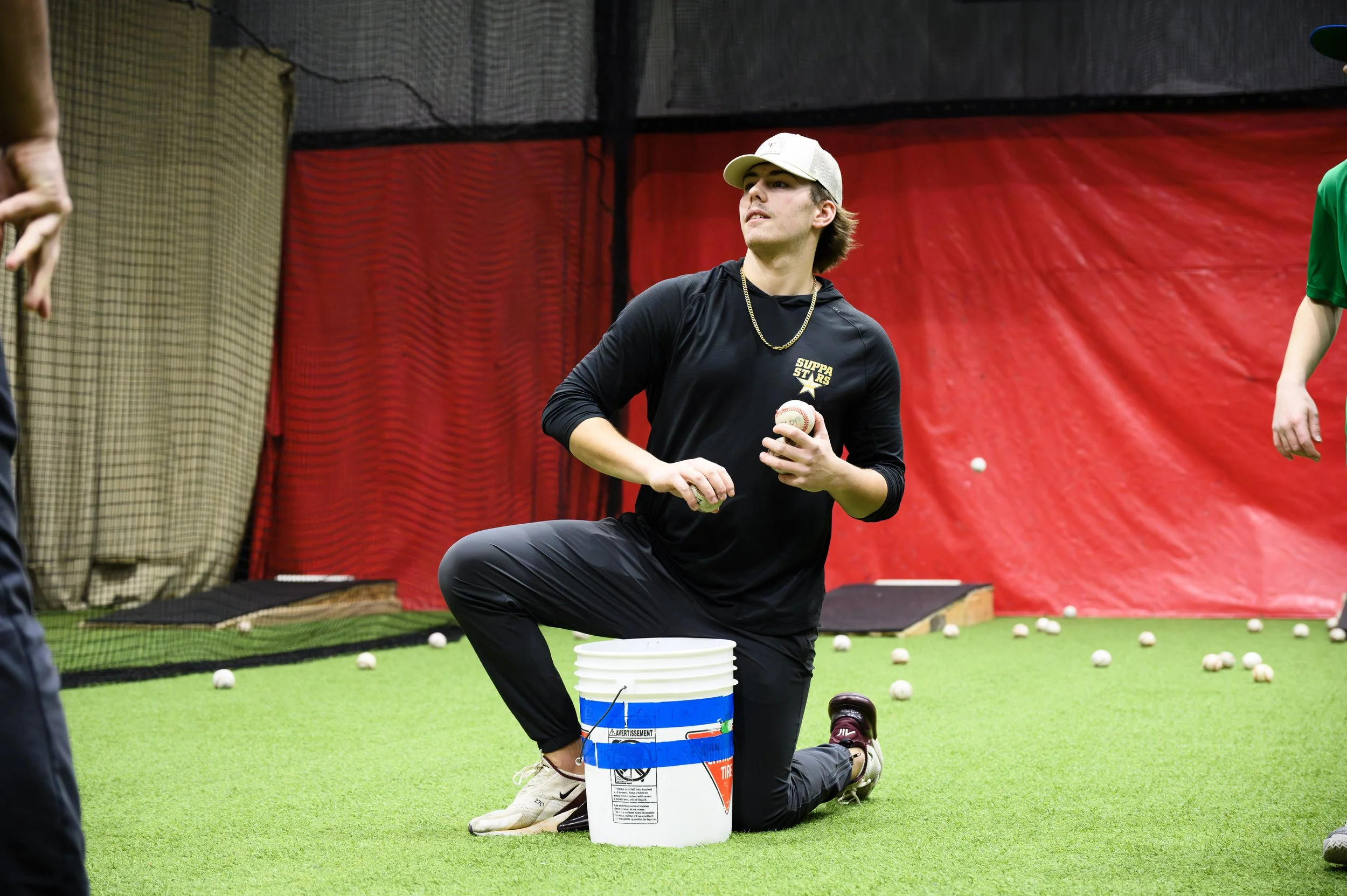 A young man in a black shirt, kneeling on one knee, holding a baseball in his right hand. He is at an indoor practice area, surrounded by numerous baseballs on the ground, with a red netted wall or backdrop behind him. He is wearing a white cap with 