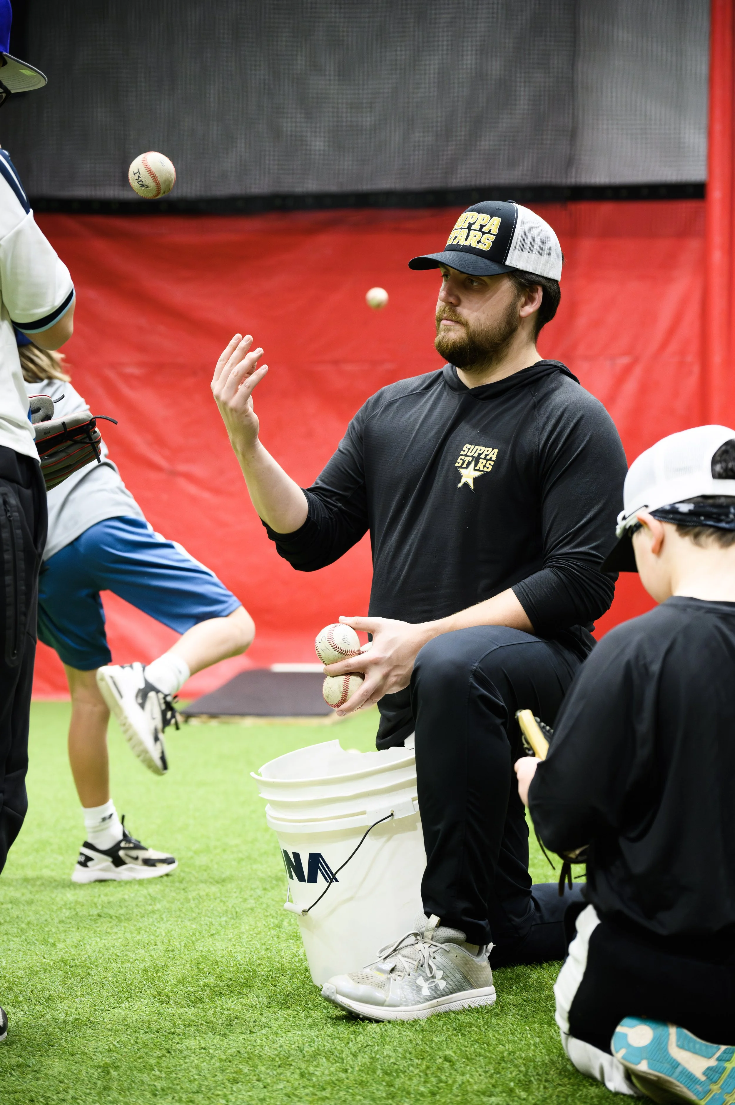 A man wearing a black hoodie and a baseball cap is holding baseballs and sitting on the ground inside an indoor baseball training facility. There are children around him, some holding baseballs, with a red wall and a large digital screen in the backg