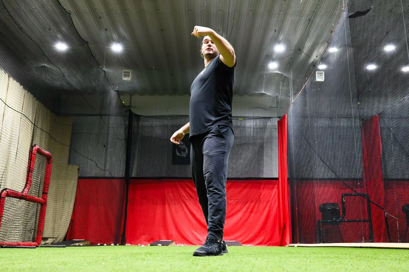 A man in black athletic clothing standing inside a batting cage with artificial turf, black netting, and red padding on the walls, preparing to hit a baseball.