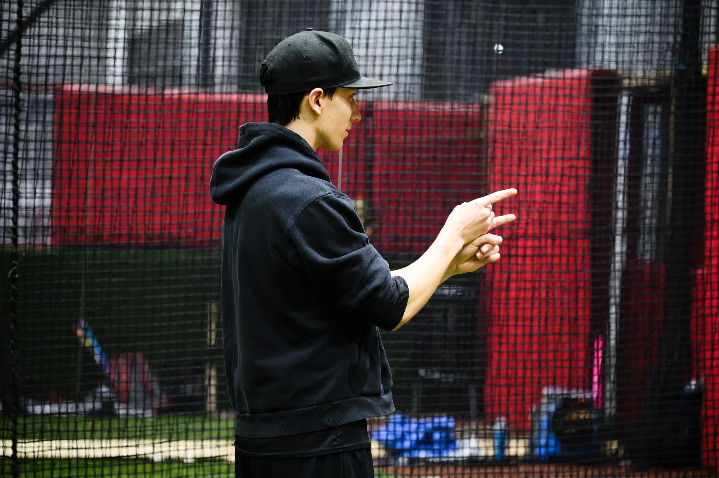 A young man in a black hoodie and black cap standing near a sports net at a baseball or softball field, with a red fence in the background.
