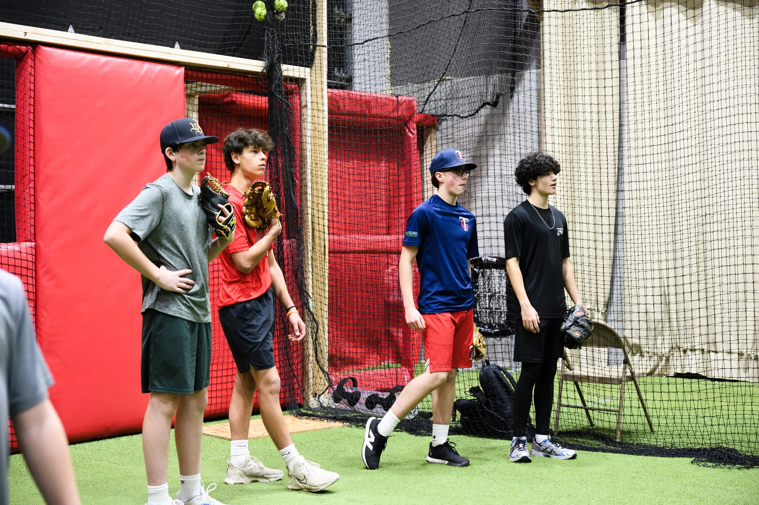 Five young boys standing in line, wearing baseball caps, holding gloves, in an indoor baseball practice area with netting behind them.