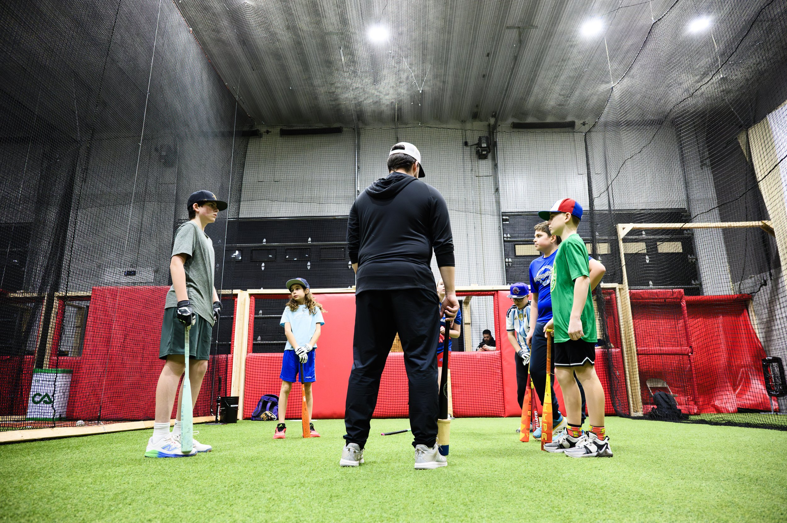 Indoor baseball or softball training session with young players and coach, a batting cage enclosed by netting, artificial grass floor, and overhead lighting.
