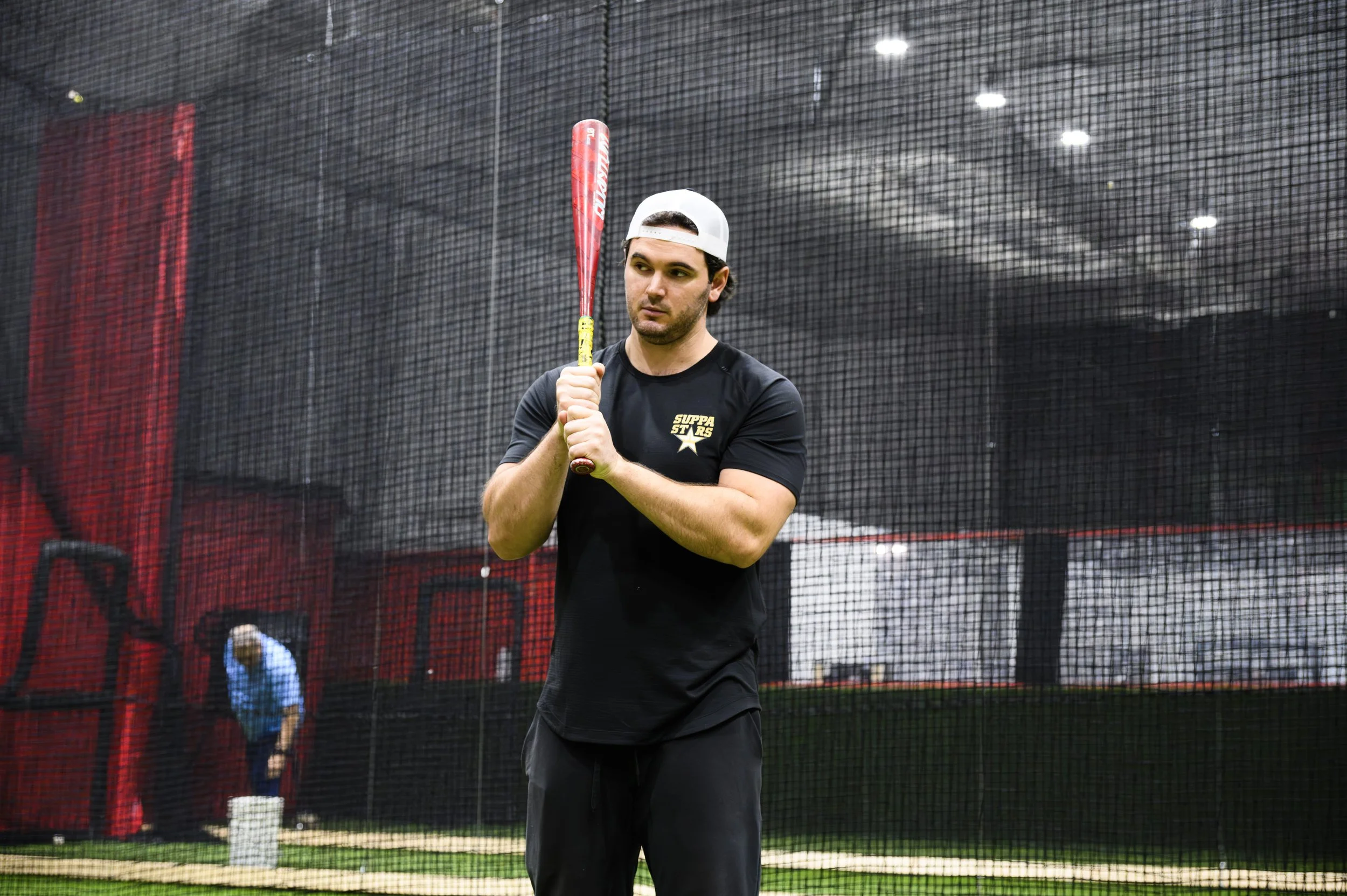 A man in a black T-shirt and white baseball cap holding a red baseball bat inside an indoor batting cage, with a person in blue and black clothing and a baseball glove in the background.