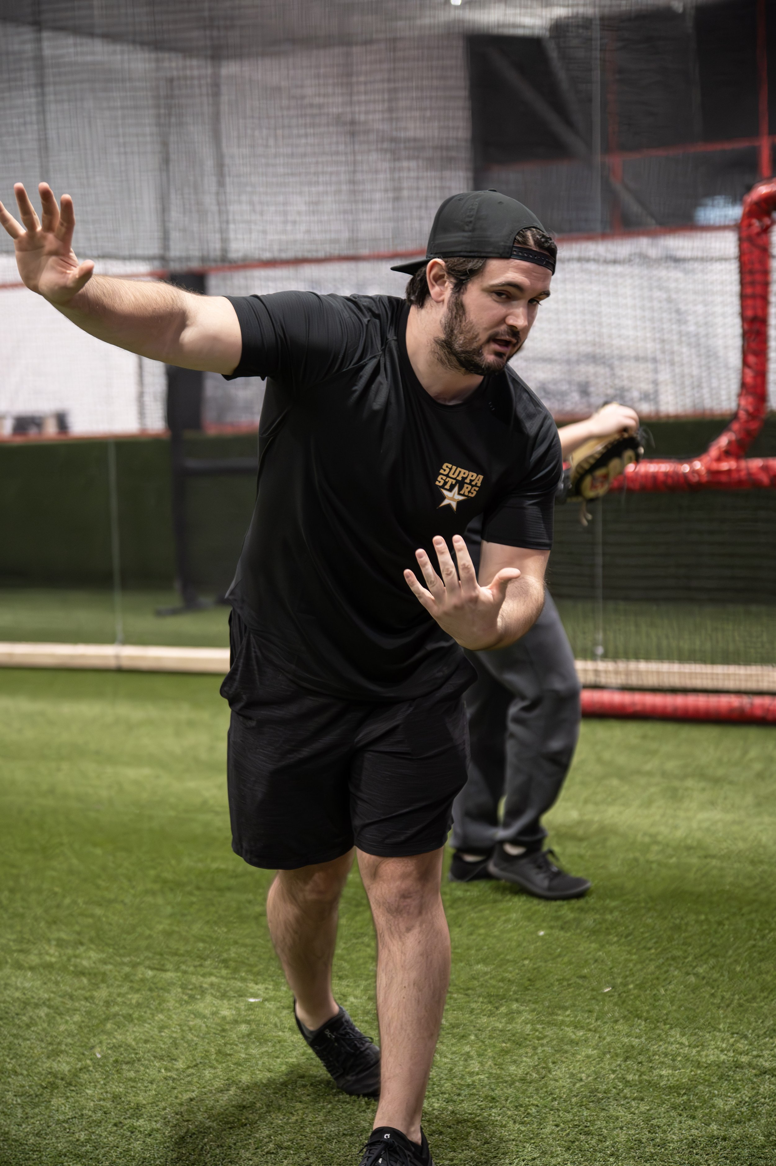 A man in a black t-shirt and shorts practicing baseball at an indoor field. He is wearing a backward black cap and has a beard. Other individuals are in the background, with one person holding a baseball glove. The field has green turf and a netted w