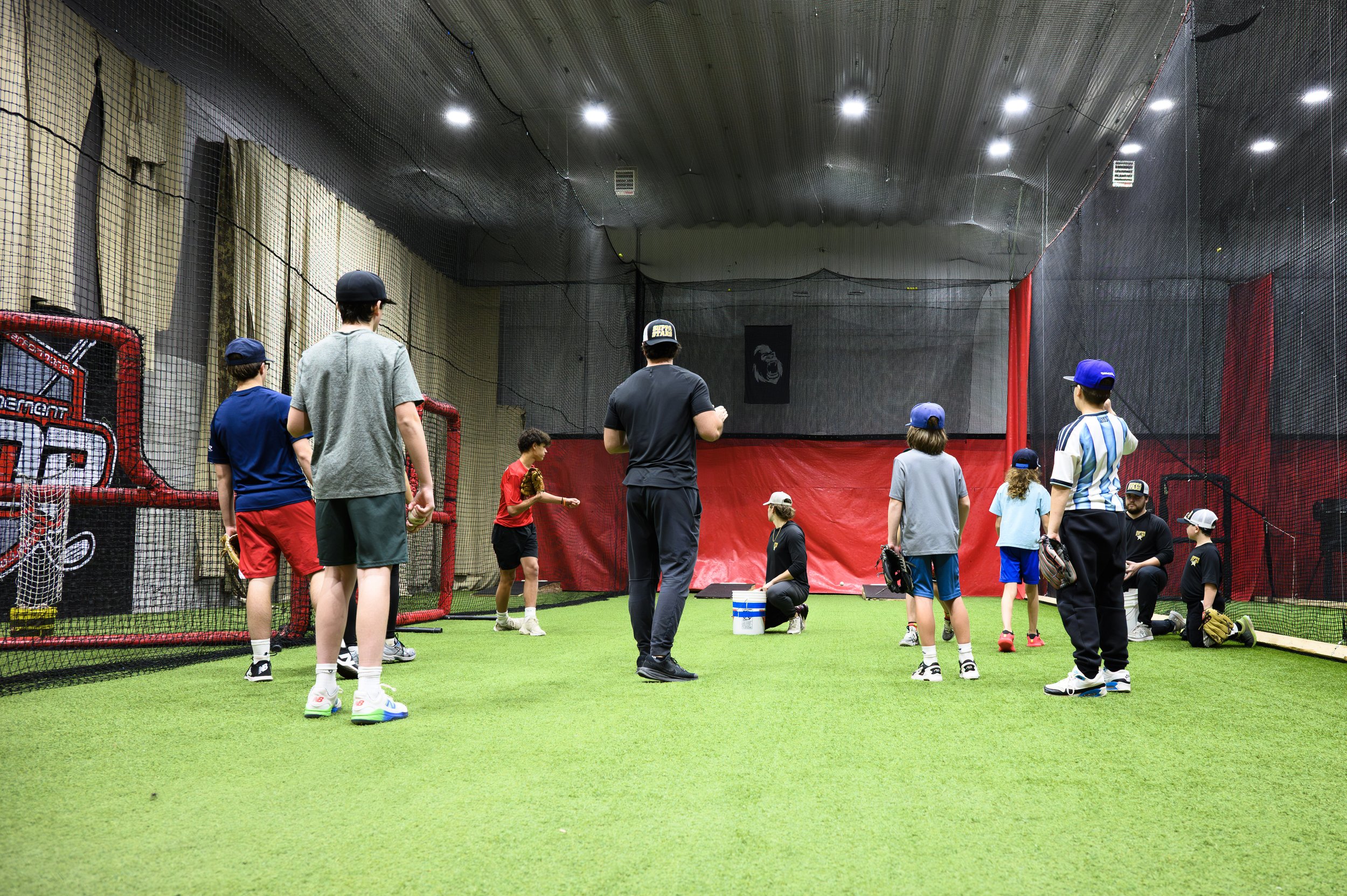 Children and adults in an indoor baseball training facility with batting cages, some children are holding gloves and baseballs, and some are sitting or standing.