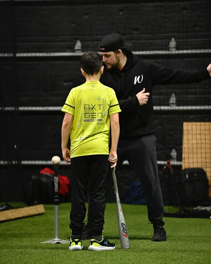 An adult male coach instructs a young baseball player during a practice or lesson indoors, with the player holding a baseball bat and wearing a yellow jersey, standing on artificial turf with baseball equipment and bags in the background.