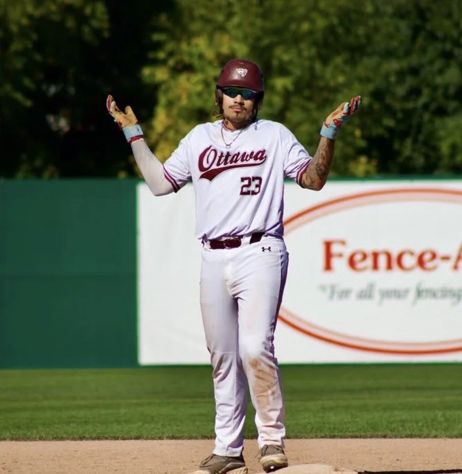 A person wearing a baseball uniform with 'Ottawa' and the number 23, standing on a baseball field with arms raised, wearing a maroon helmet and sunglasses.