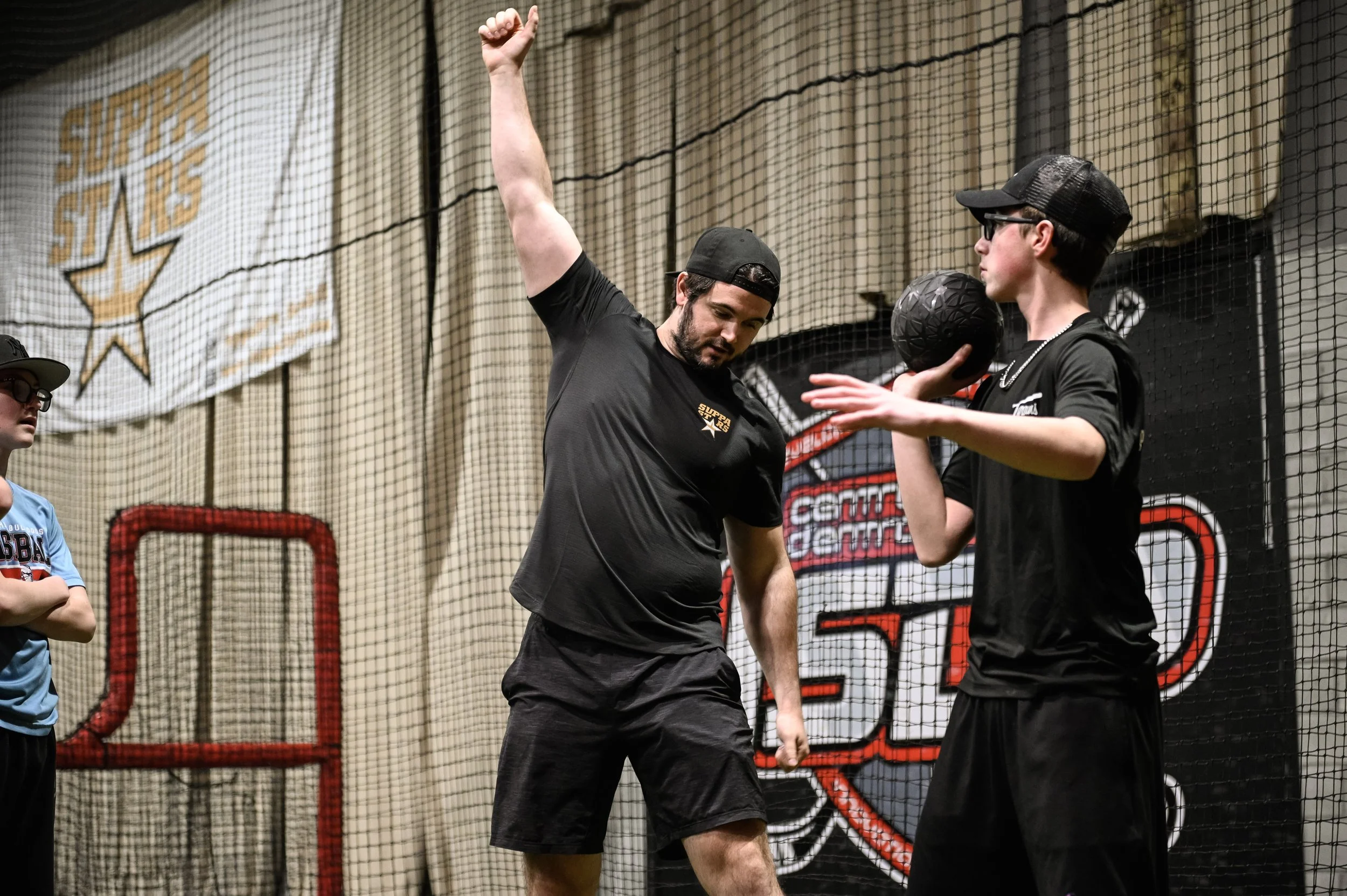 Two young men in black sportswear holding a black medicine ball during an indoor sports practice. One is raising his arm and the other is wearing a baseball cap and glasses. A girl in a blue shirt and cap observes nearby. Behind them, sports equipmen
