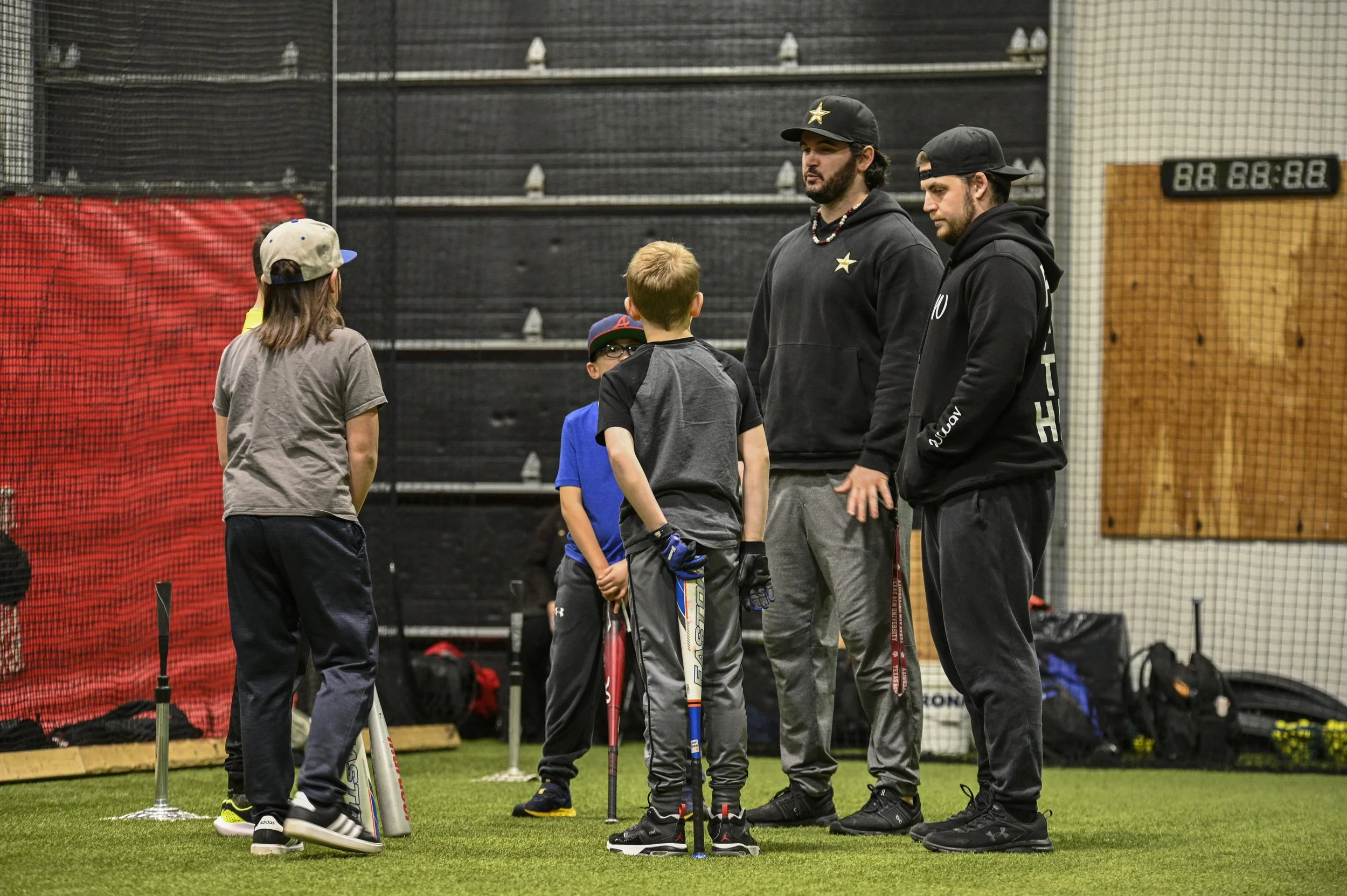 Two adult men and four children in a sports training facility, standing on artificial turf, surrounded by sports equipment and a digital timer on the wall.