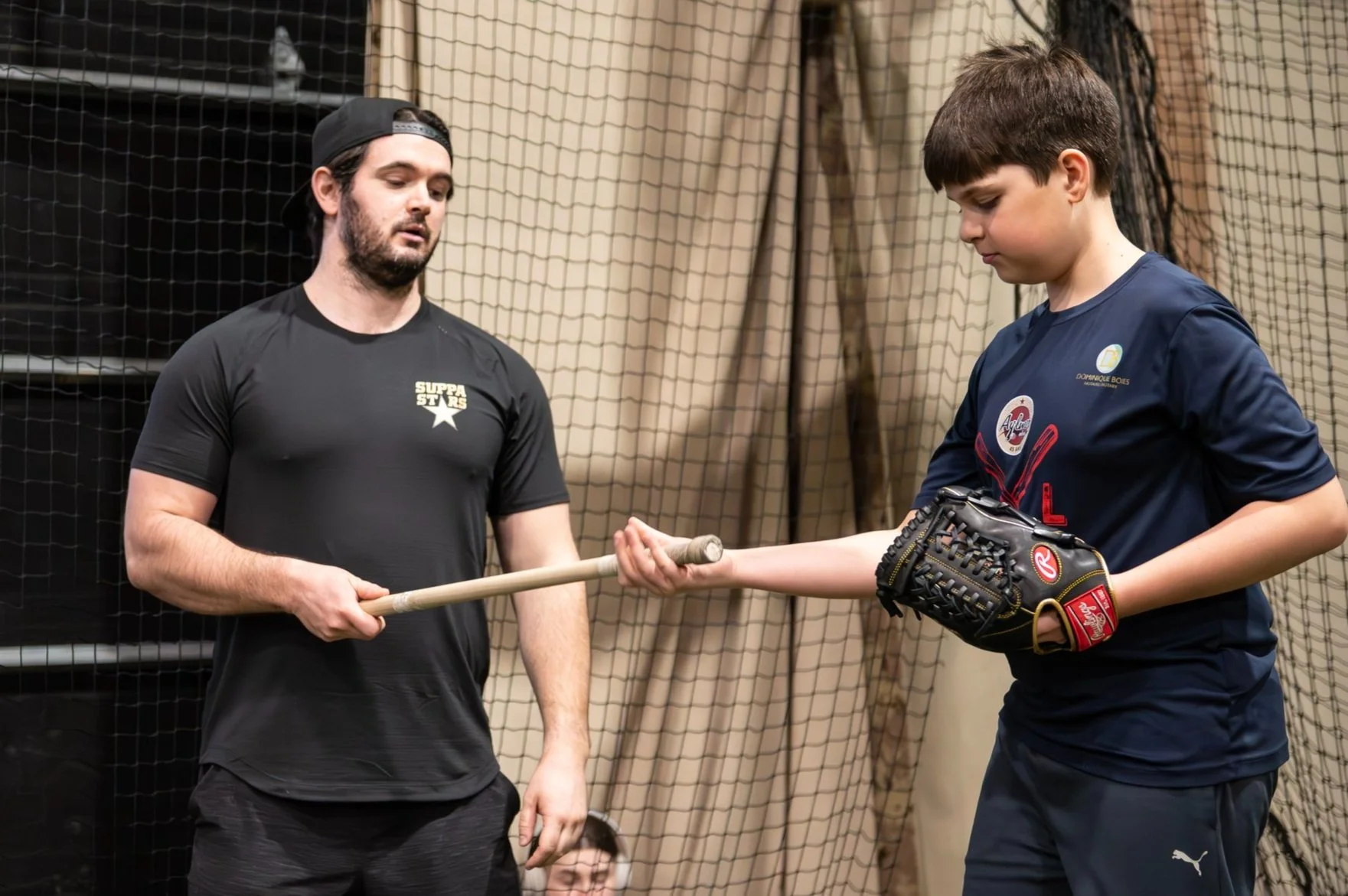 A man wearing a black t-shirt and a black cap coaches a young boy in a baseball glove, holding a baseball bat, inside a sports facility with netting in the background.