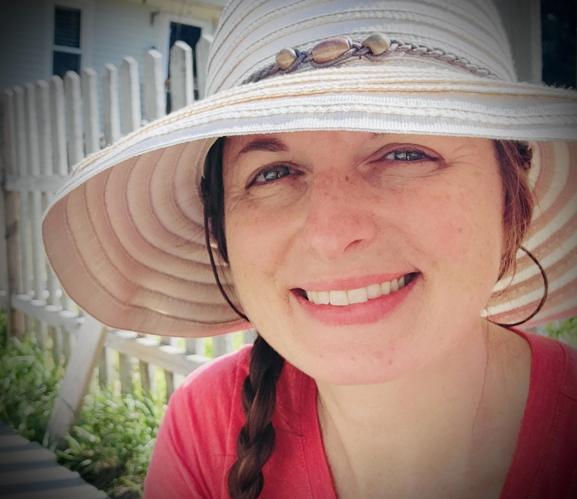 Close-up of a smiling woman wearing a large sun hat with shell decorations, outdoors near a white picket fence, and a house in the background.
