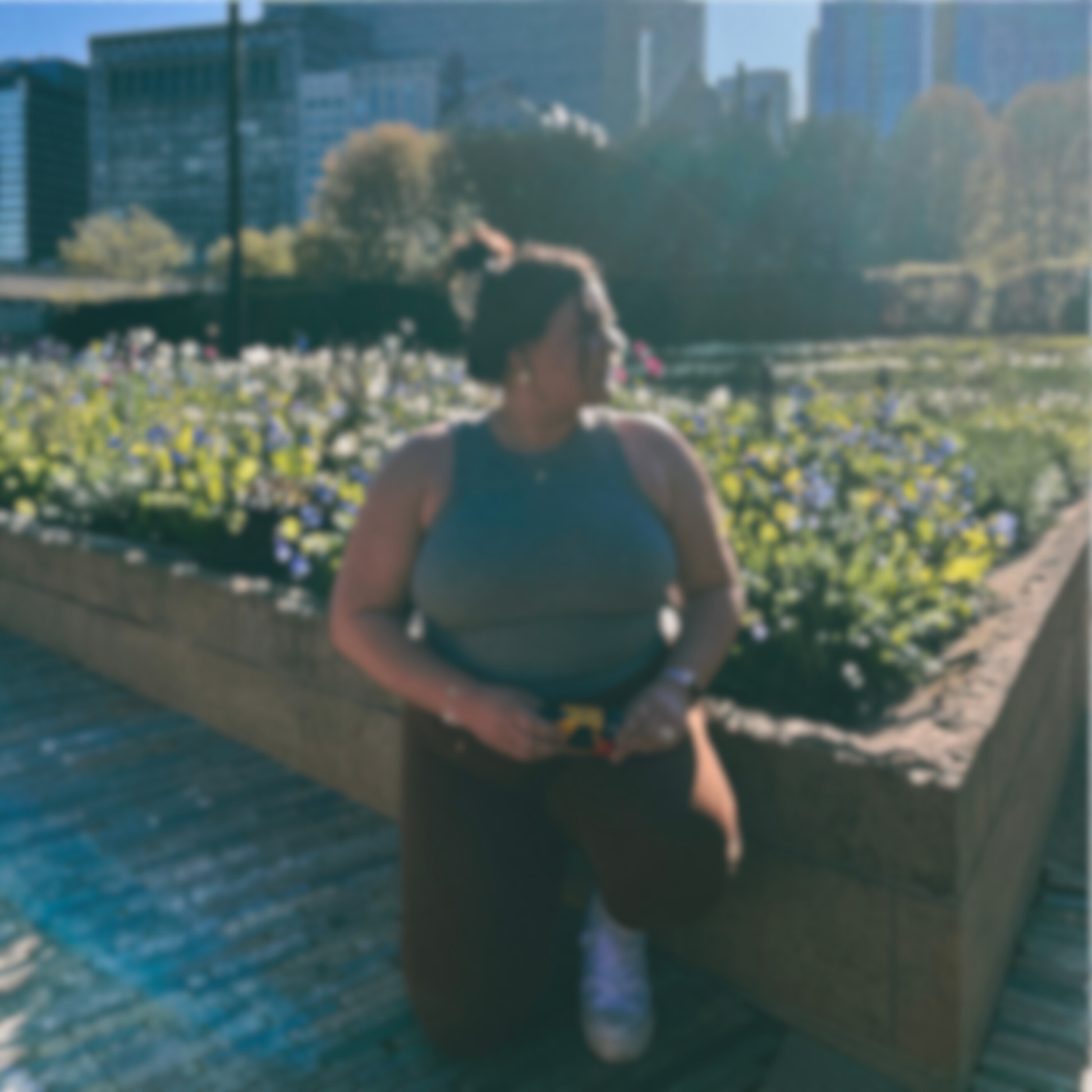 A woman in athletic clothing stands beside a flower bed in an urban park, with city buildings in the background.