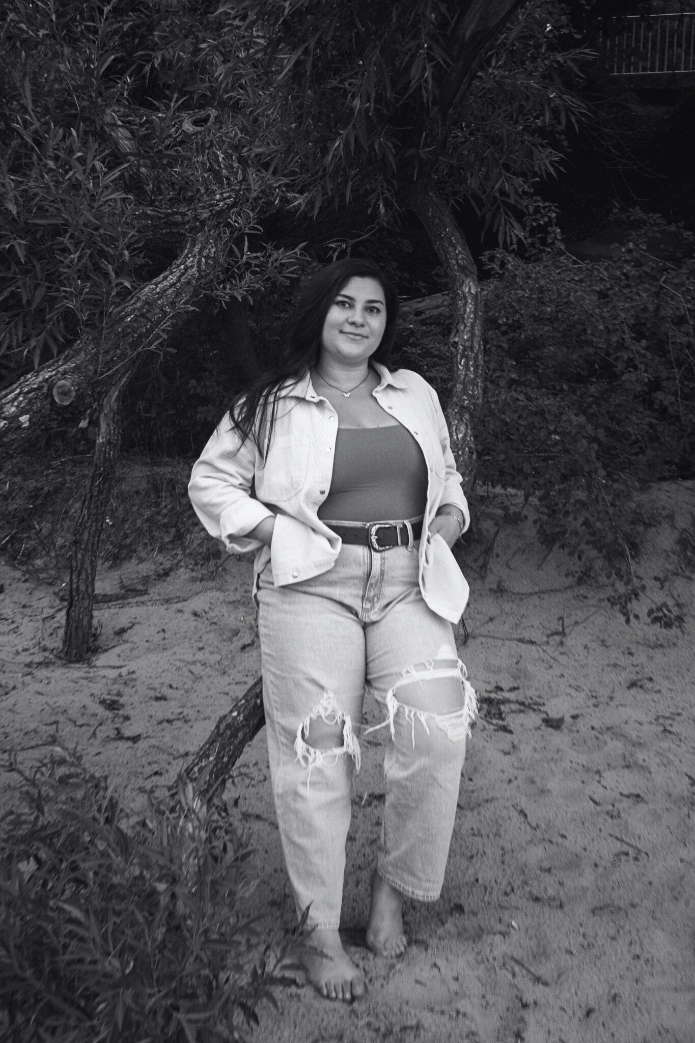 Jessica standing barefoot on sandy ground next to a tree with dense foliage behind her, wearing ripped jeans, a tank top, and a jacket, in black and white.
