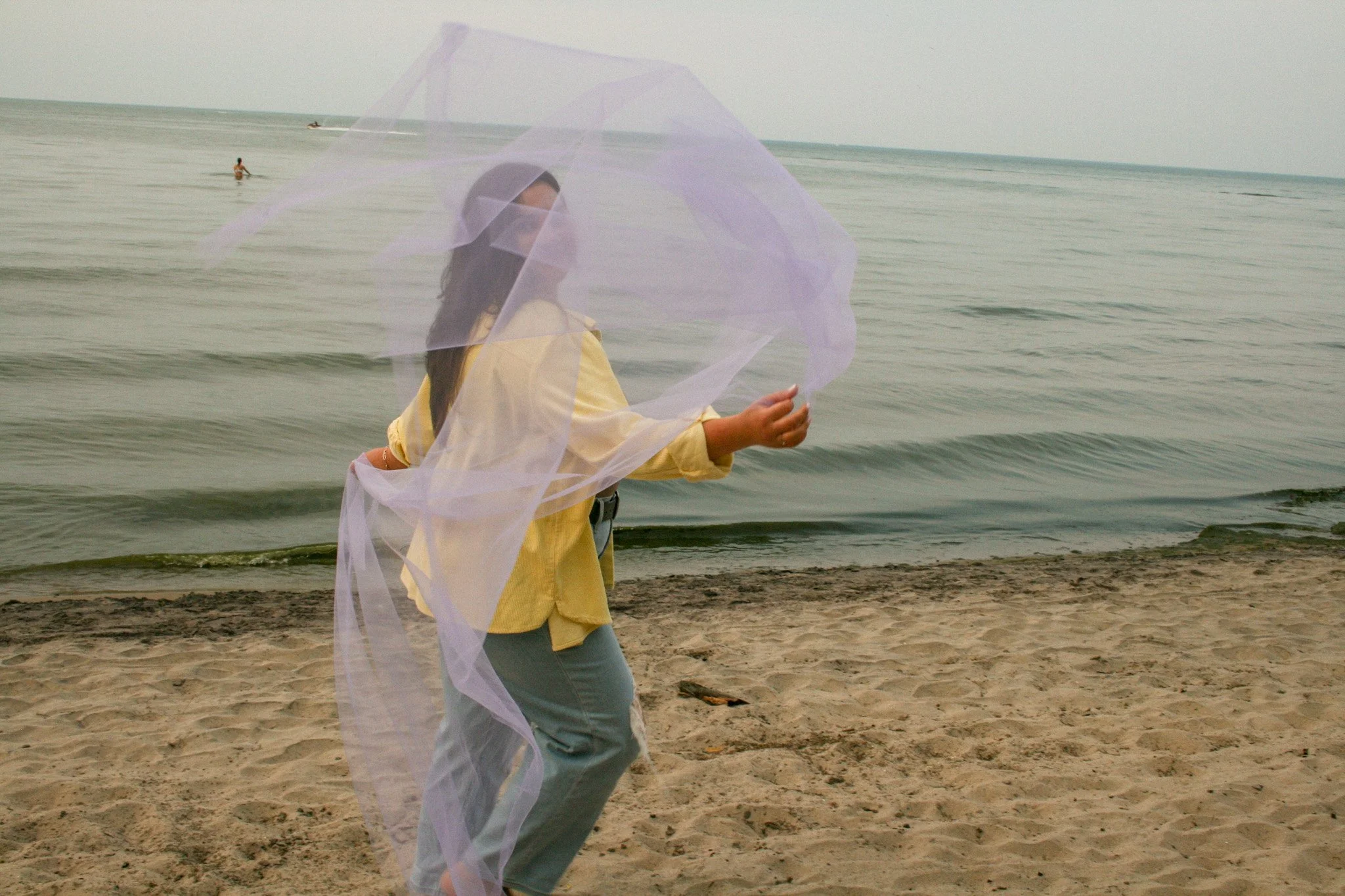 Woman in yellow shirt and gray shorts on a sandy beach holding a transparent purple umbrella, with the ocean and a person swimming in the background.