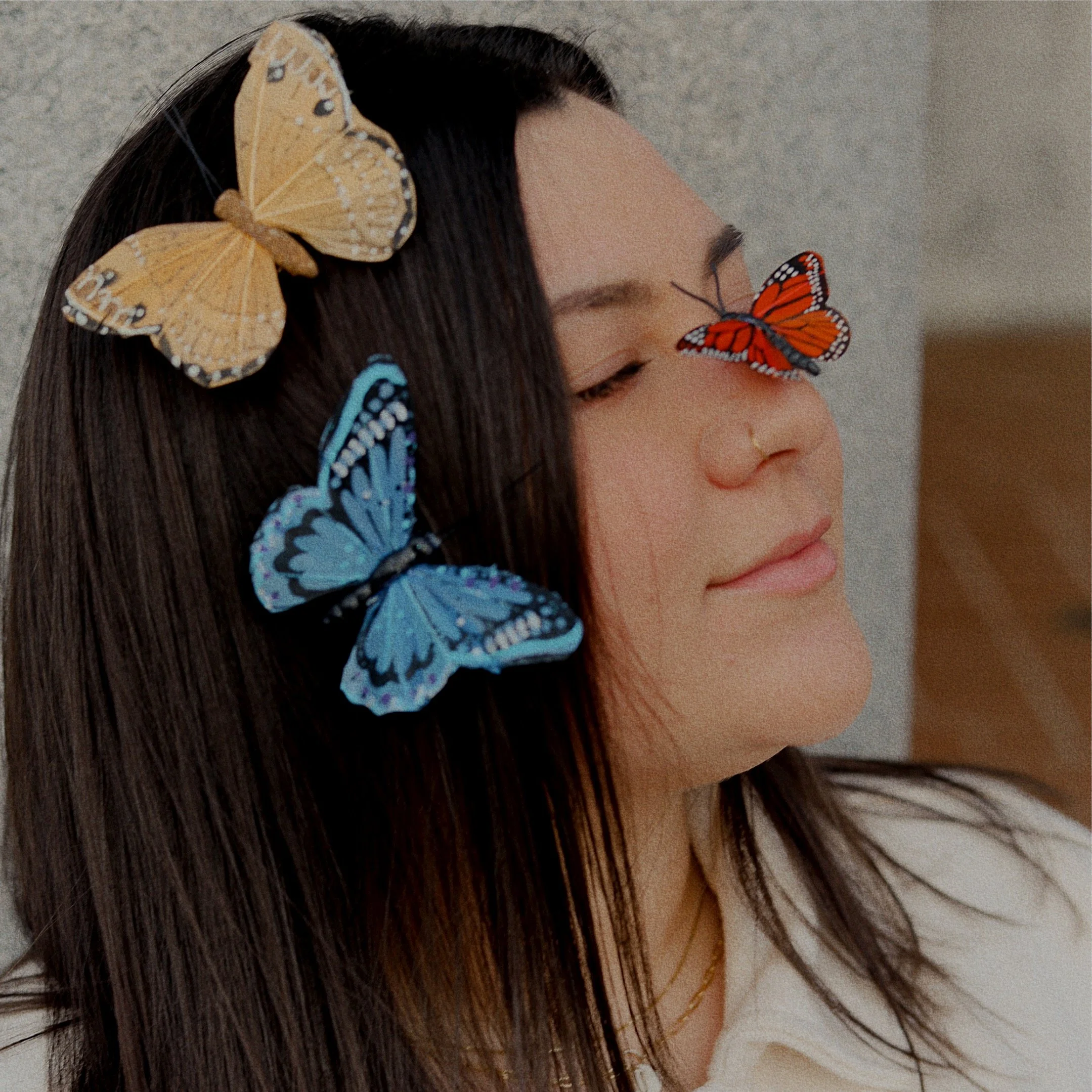 A woman with long dark hair has her eyes closed and a peaceful expression, with three colorful butterflies resting on her face and hair.