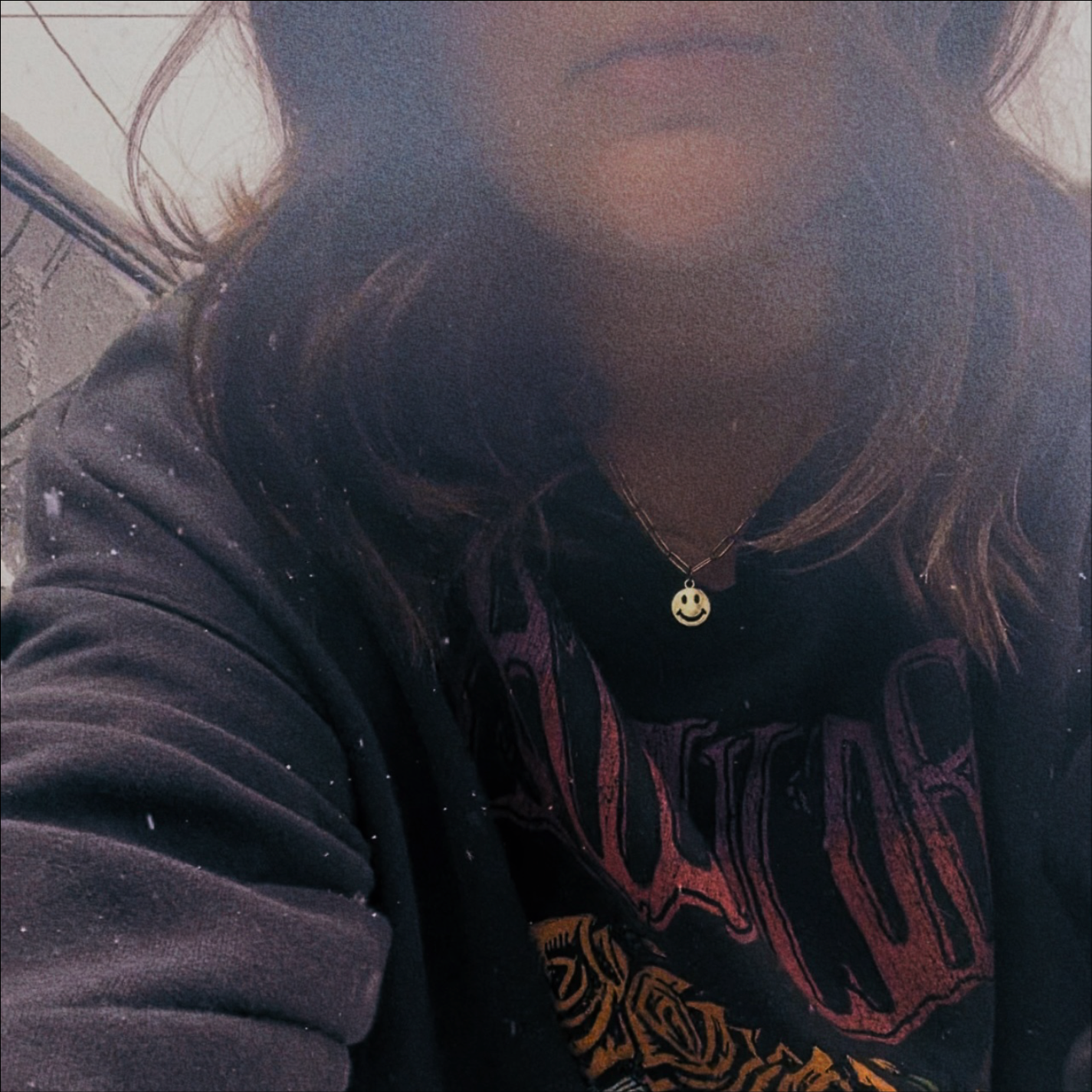 A person with dark, shoulder-length hair wearing a black hoodie and a necklace with a smiley face pendant, standing outdoors near a brick wall.