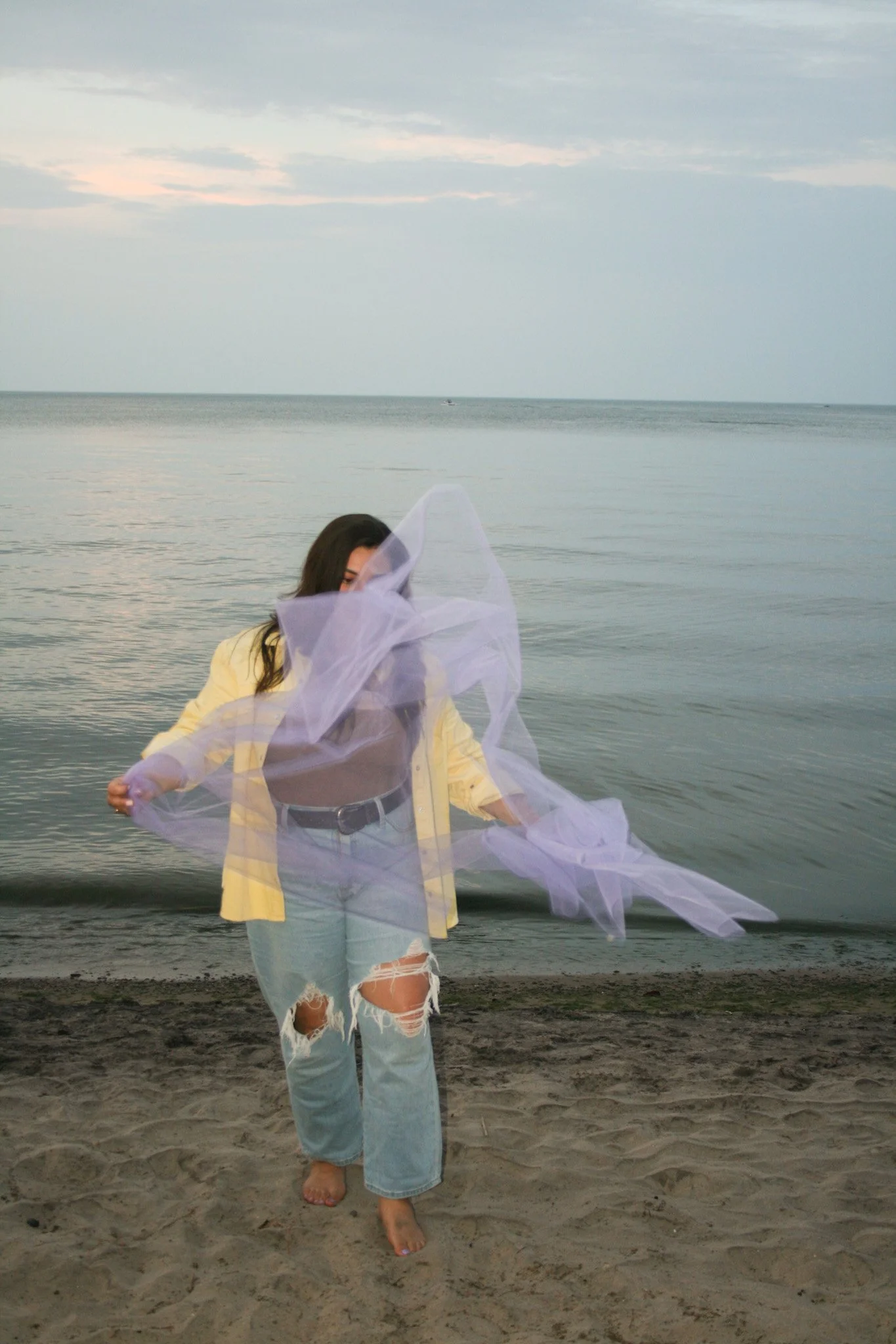 Woman walking along the beach with a semi-transparent fabric flowing in the wind, ocean and cloudy sky in the background.