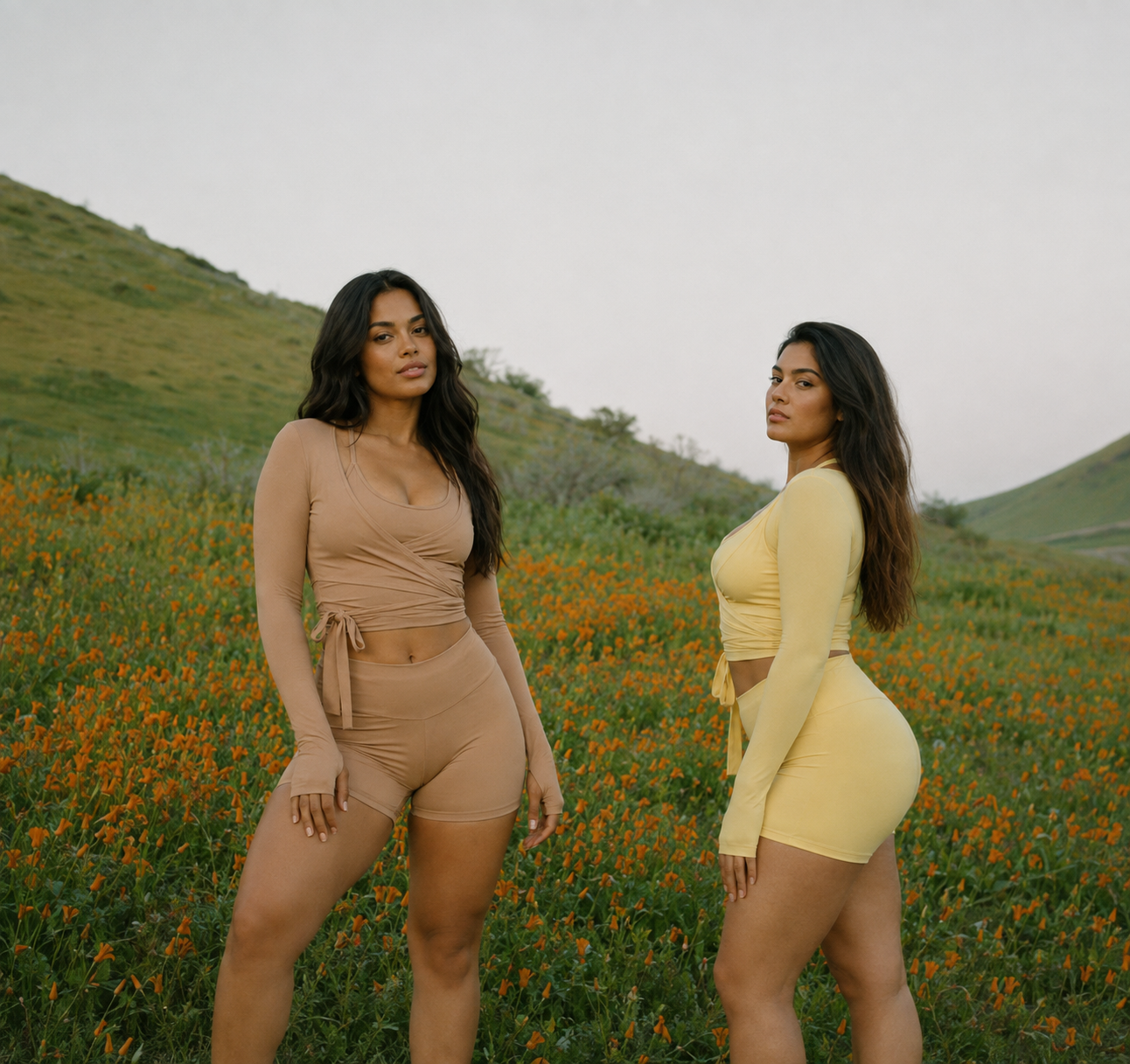 Two women standing in a field of orange flowers with green hills in the background, wearing beige and yellow matching outfits.