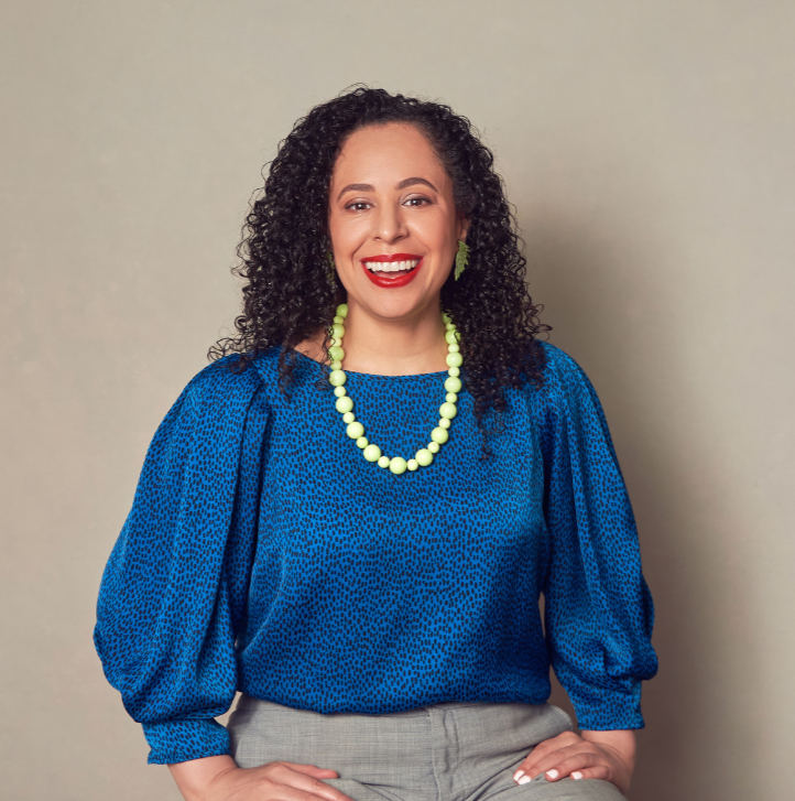 A woman with curly black hair, wearing a bright blue patterned blouse, light gray pants, and accessories including green earrings and a matching green beaded necklace, smiling against a plain background.
