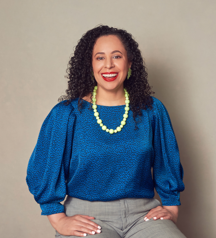 A woman with curly dark hair wearing a bright blue blouse, beige pants, and green jewelry, smiling while sitting against a plain background offering anxiety therapy for in NYC.