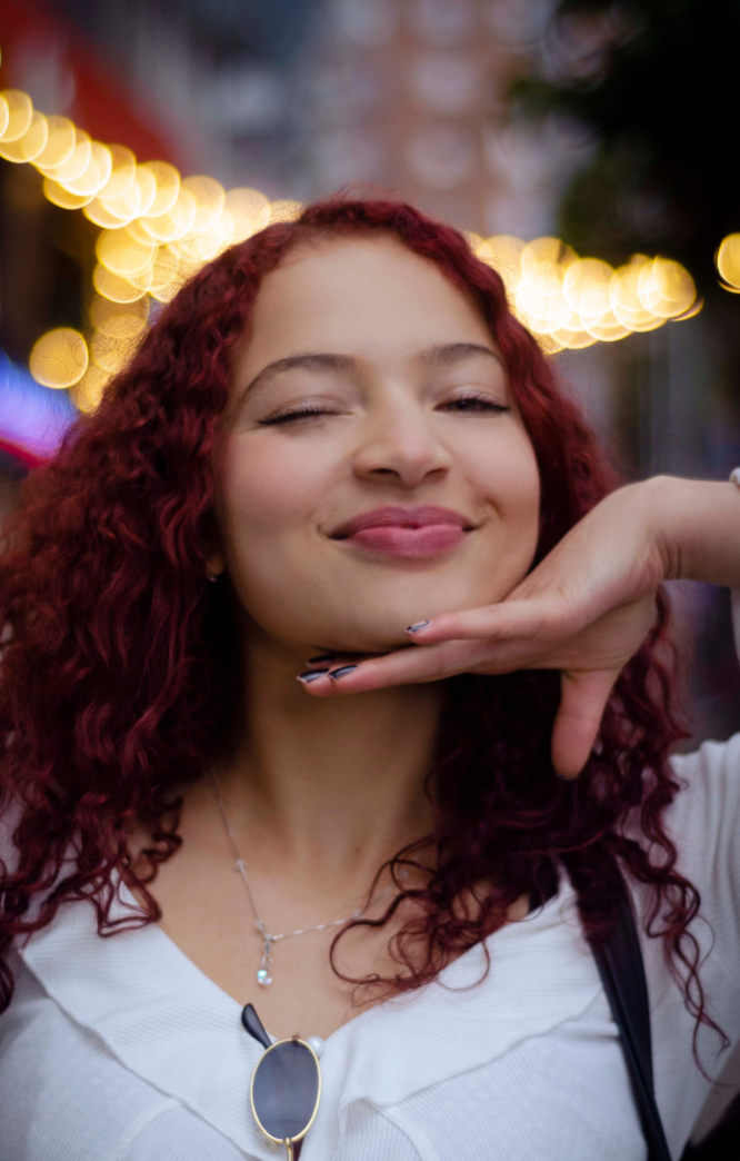 A young woman with curly red hair smiling and posing with her hand under her chin, outdoors with blurred string lights in the background.