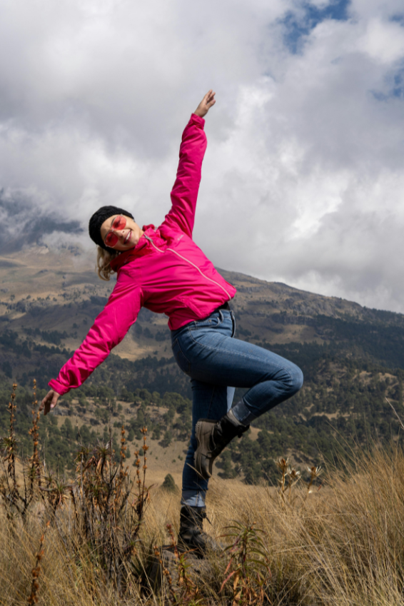 Woman in pink jacket and sunglasses balancing on one leg in a grassy field with mountains and cloudy sky in the background.