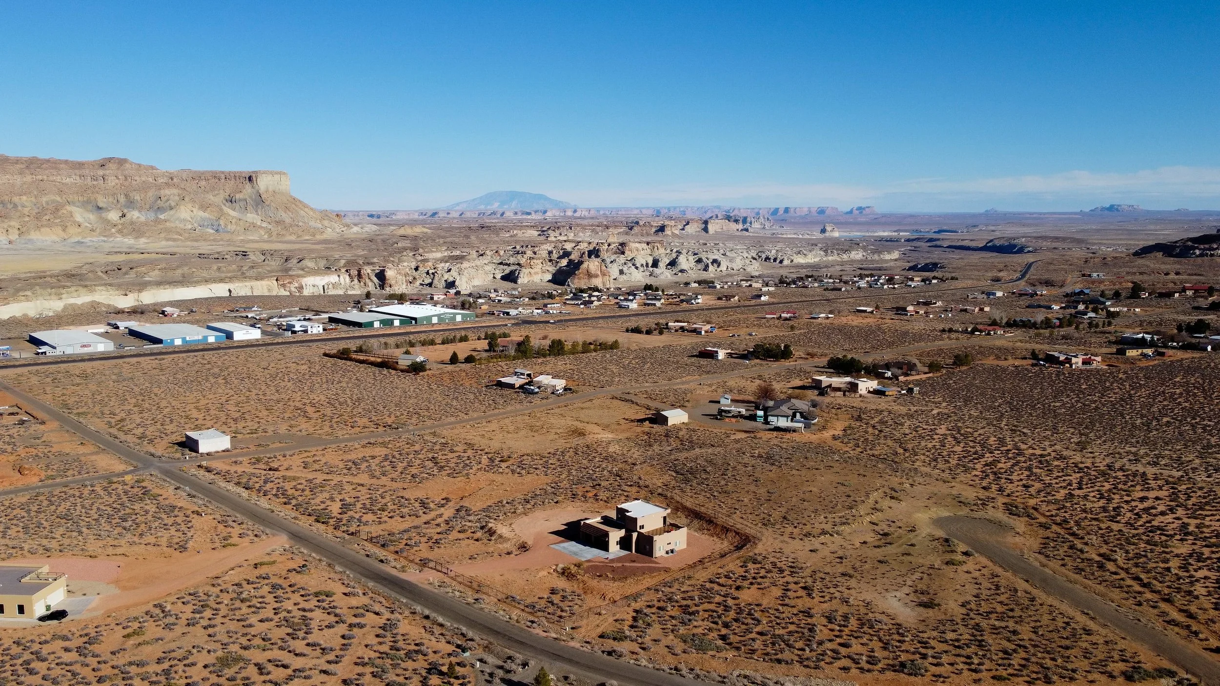 Aerial view of a desert landscape with scattered homes, roads, and industrial buildings, with rocky mesas and cliffs in the background.