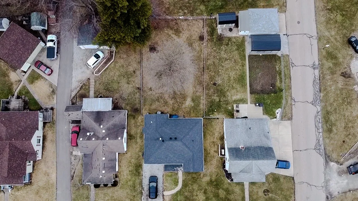 Aerial view of a neighborhood with houses, cars, lawns, and empty backyard lots.