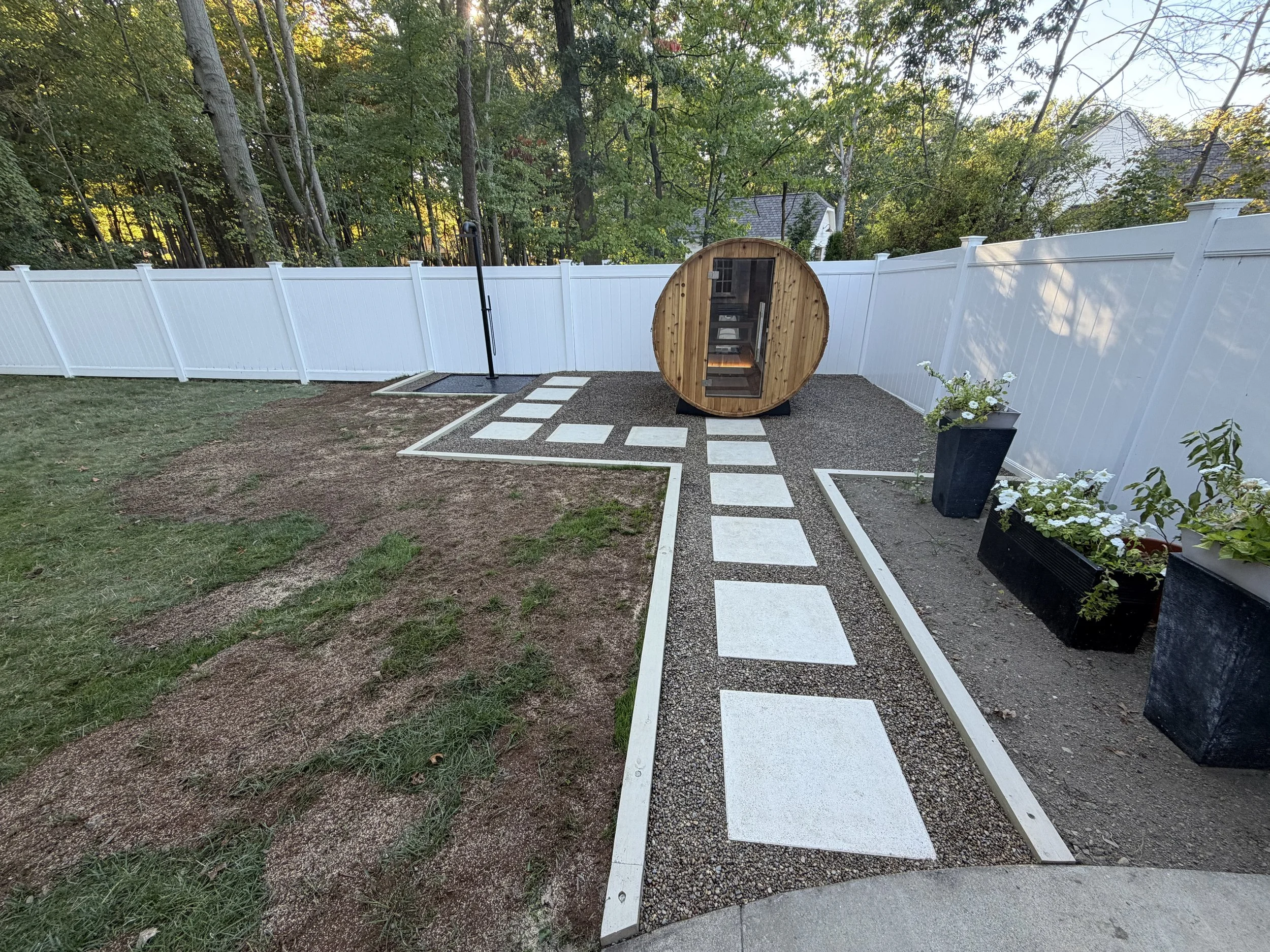 Backyard with a white fence, stepping stones, a green lawn, potted plants, and a small wooden sauna.