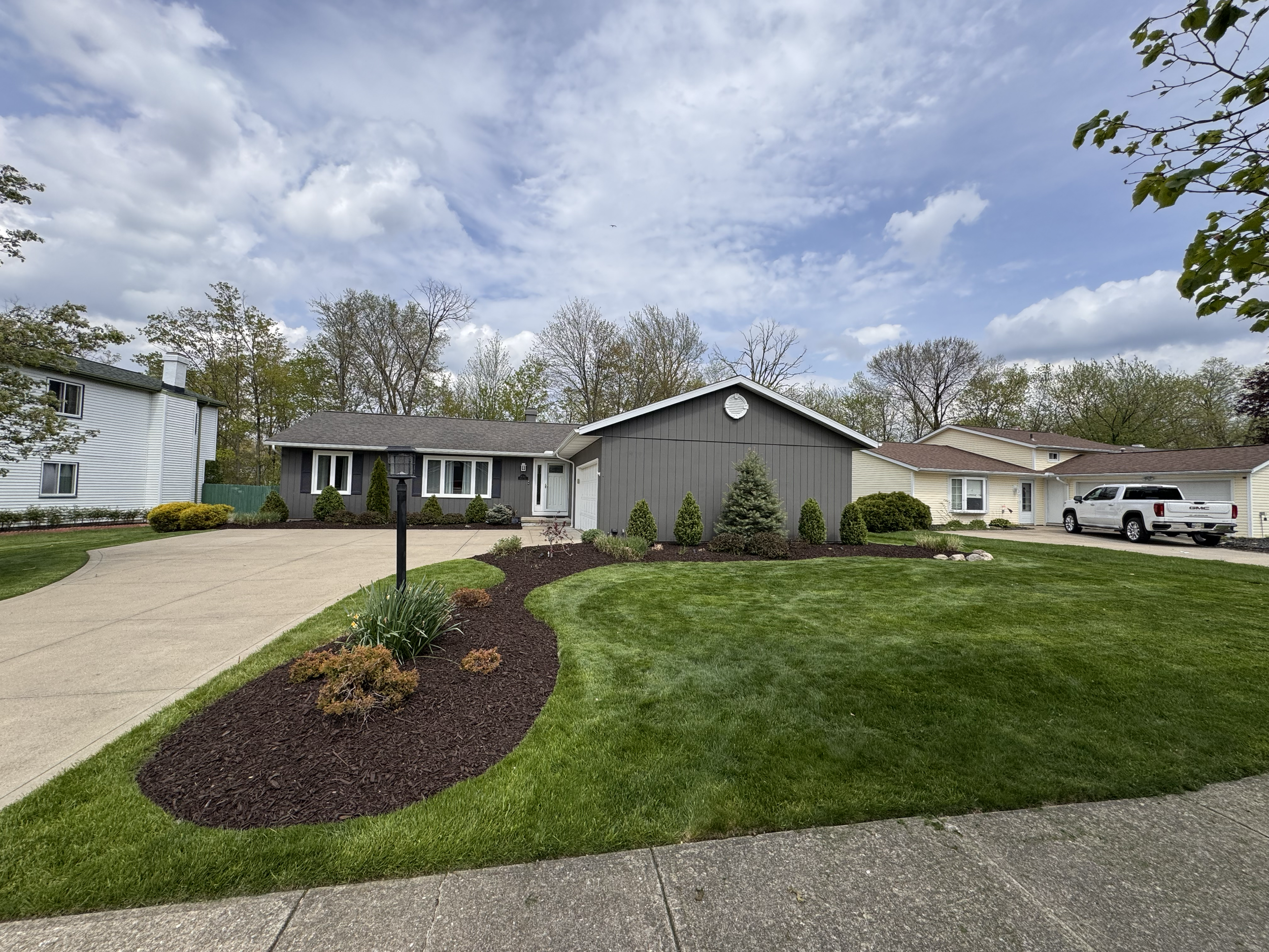 Front yard of a house with a curved driveway, green lawn, various shrubs, small trees, and a street lamp, under a partly cloudy sky.