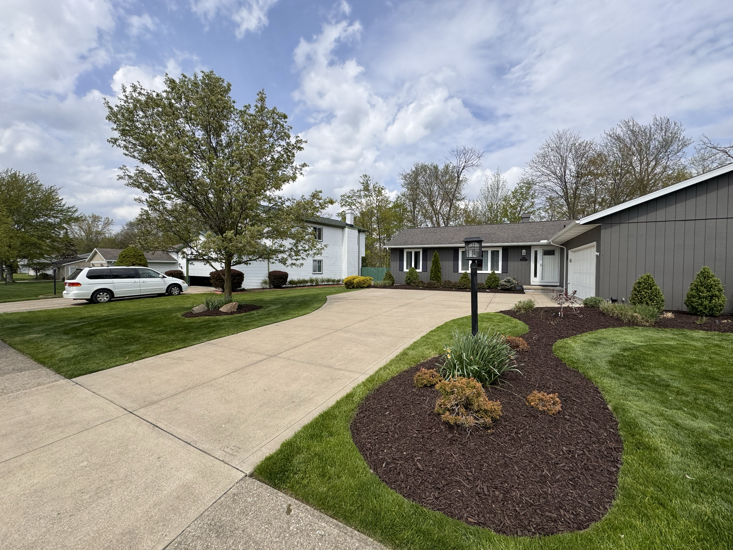 Front yard of a suburban house with a curved driveway, green lawn, landscaped flower beds, trees, and a parked white vehicle.