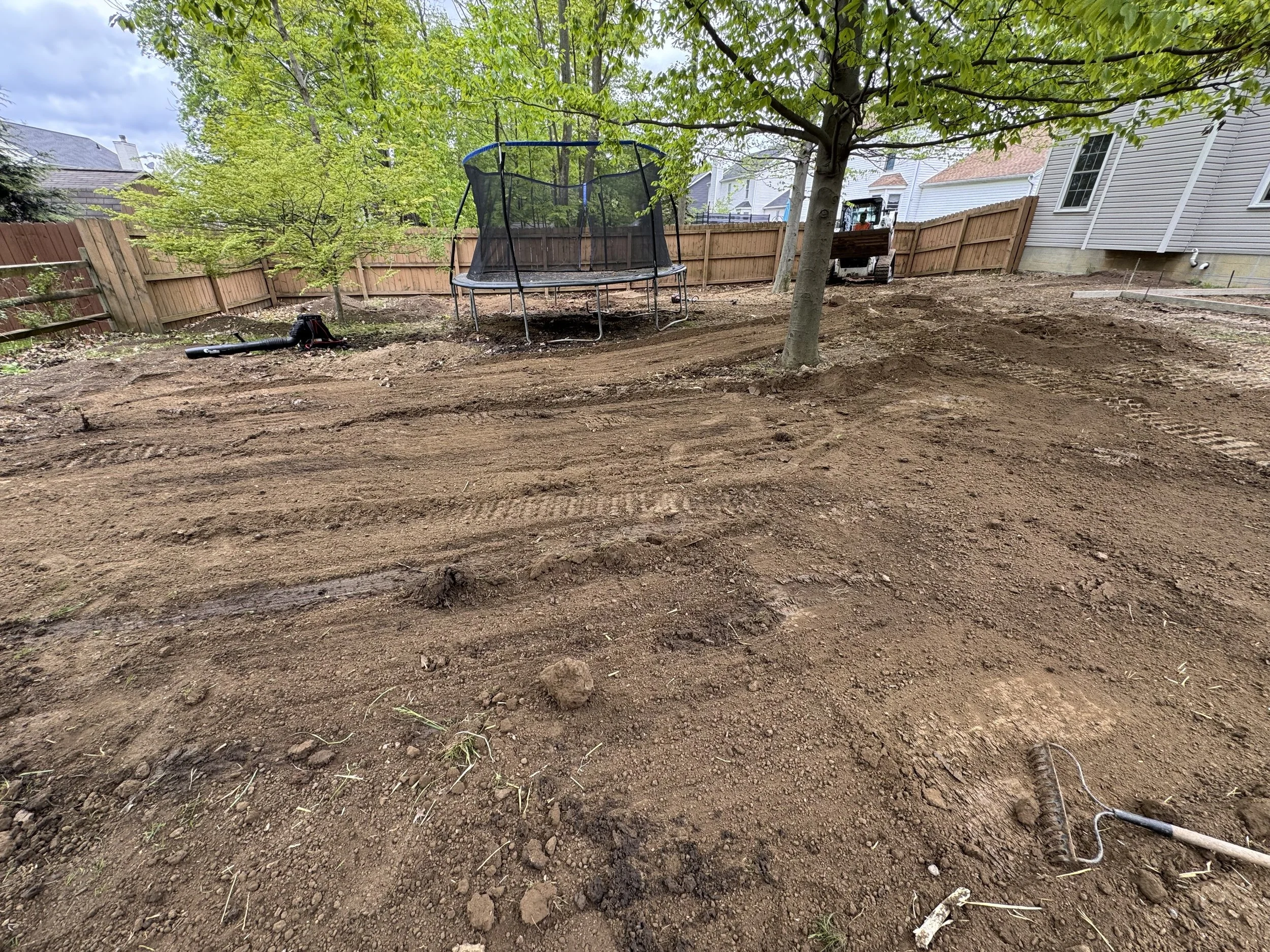Backyard with freshly tilled soil, a tree, a trampoline, a leaf blower, and a garden rake, with a wooden fence and neighboring houses in the background.