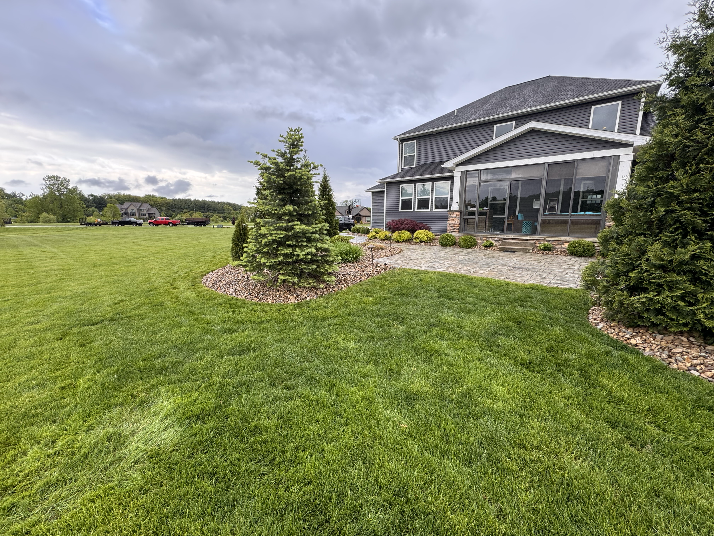 Backyard with a well-maintained lawn, small trees, shrubs, a stone pathway, and a two-story house with a screened porch under a cloudy sky.