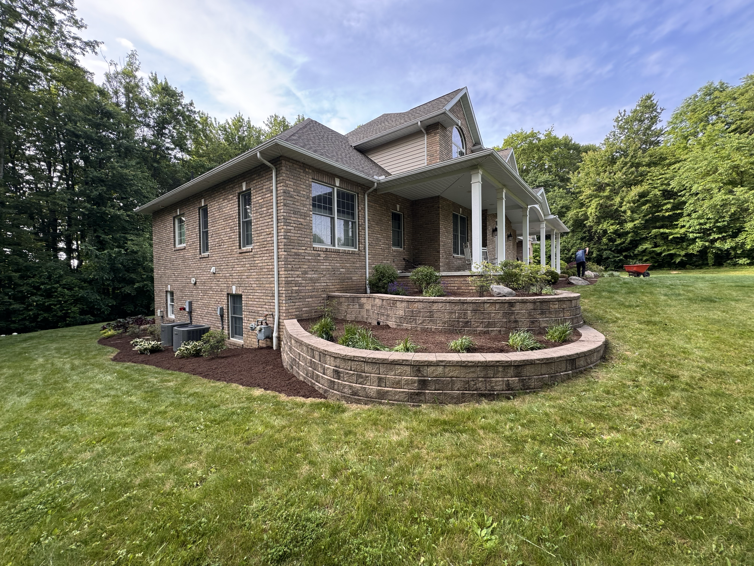Brick house with a green lawn and landscaped flowerbeds, white porch columns, and a person working in the background.