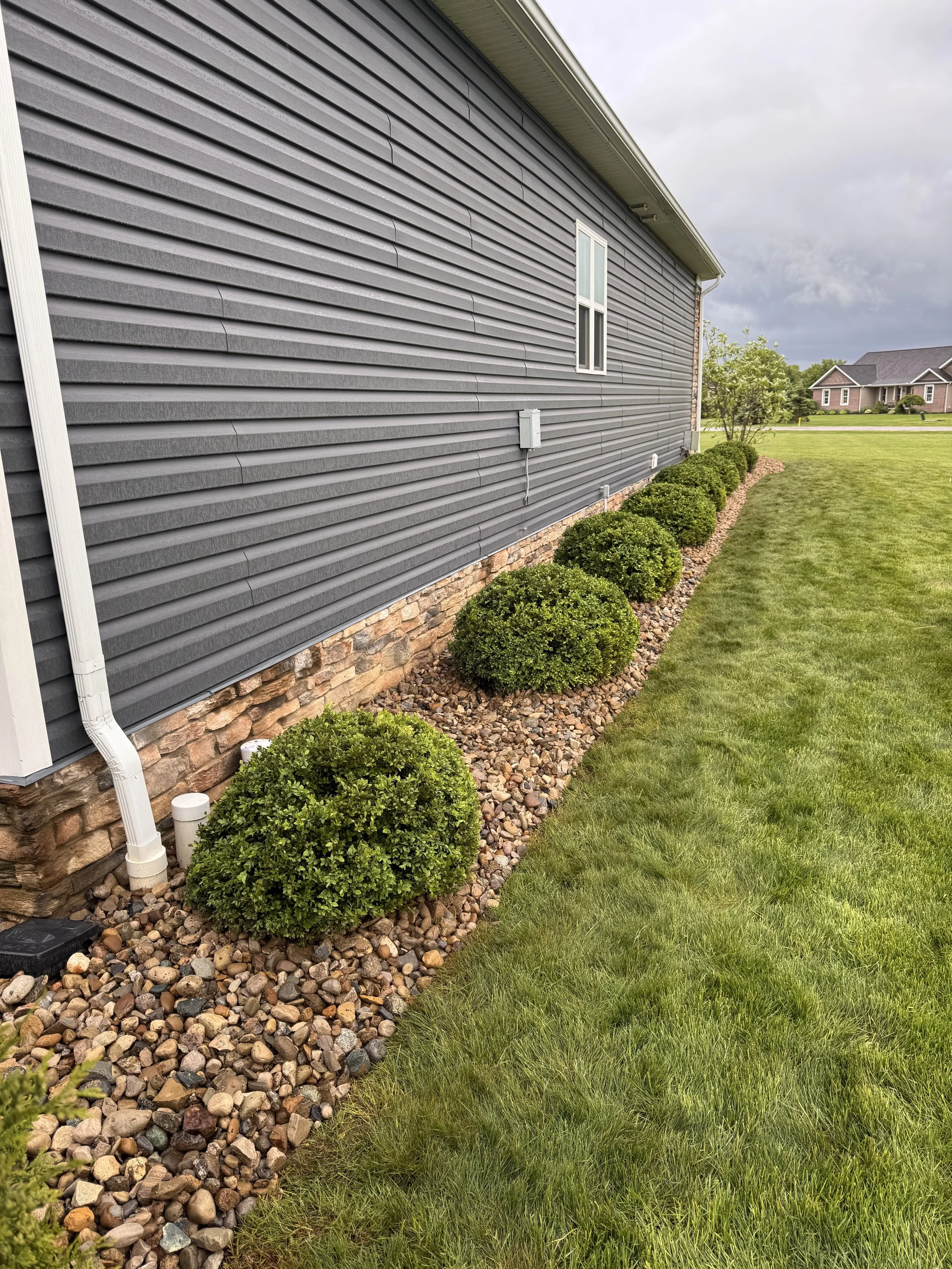 Side of a house with grey siding, a small window, a white downspout, and a bed of rounded rocks with neatly trimmed bushes along the foundation. A lawn extends in the foreground with other houses in the background.
