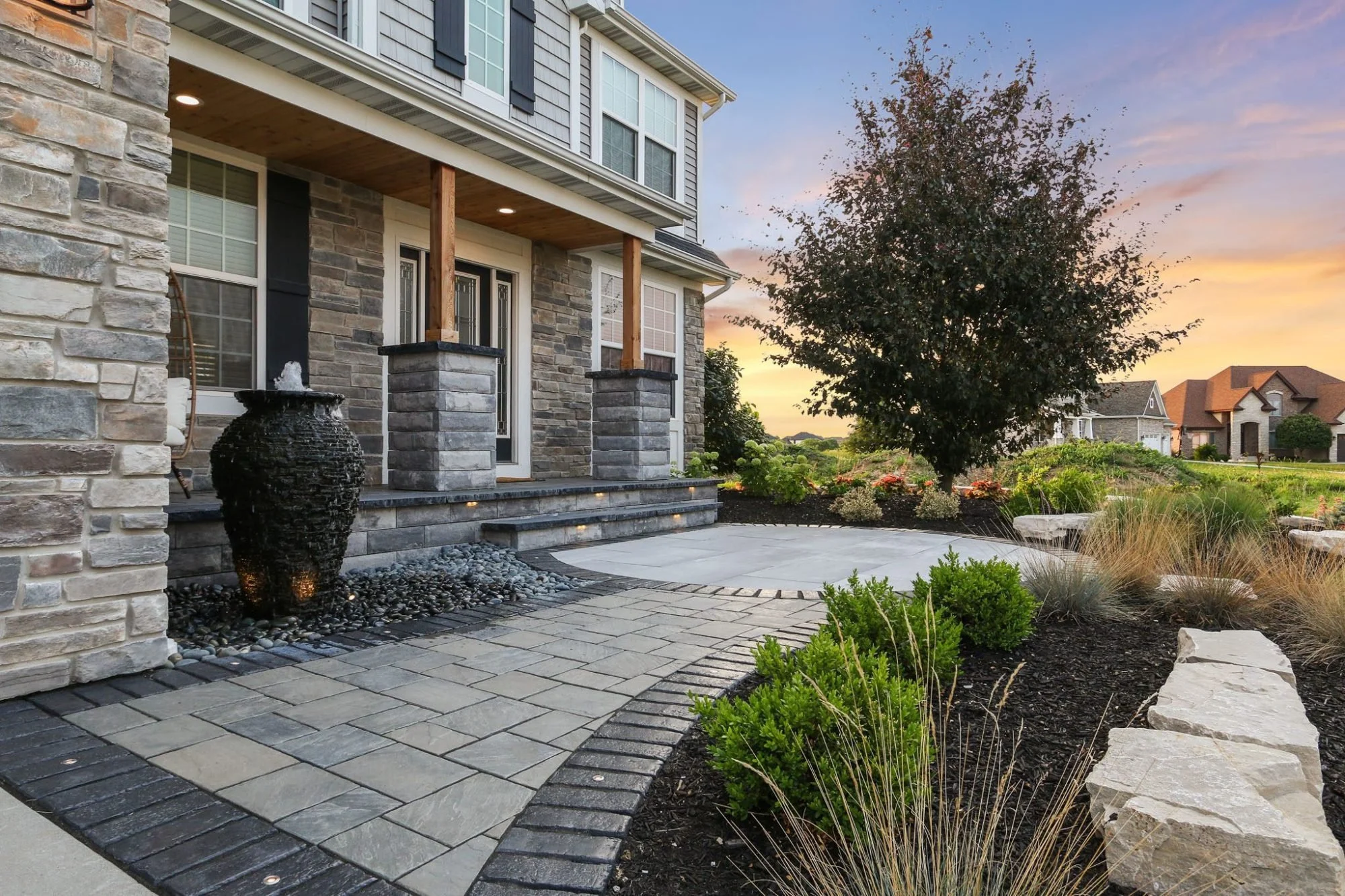 Front yard of a house with stone facade and pathway, decorative water fountain, landscaped garden, and sunset sky in the background.