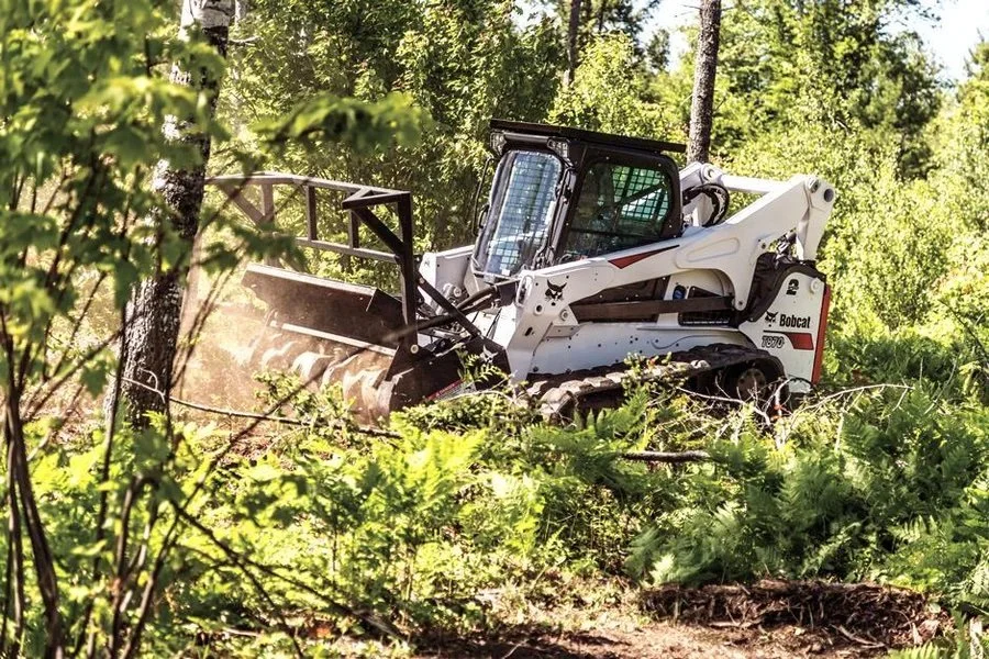 Skid steer loader with forestry attachment clearing trees in a forest.
