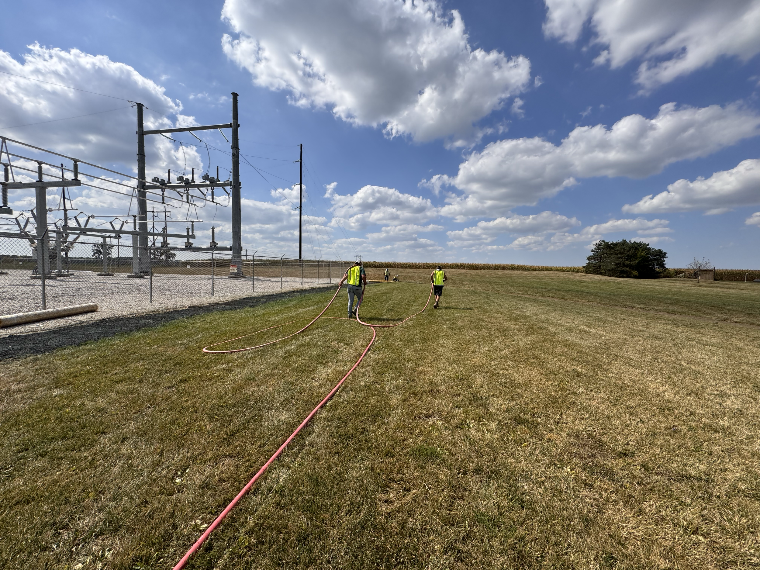 Two workers in safety vests watering or inspecting an open grassy field near electrical substation fences and power lines under a partly cloudy blue sky.