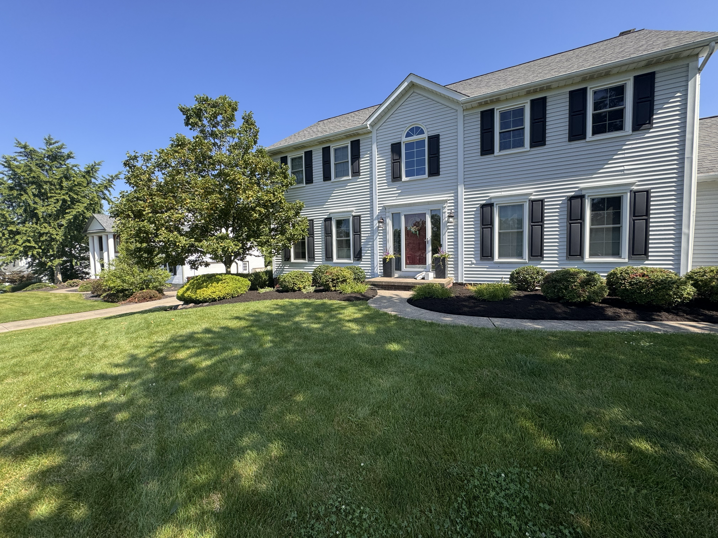 A white two-story house with black shutters and a red front door, surrounded by a well-maintained lawn and landscaped bushes, with a tree in front and a clear blue sky overhead.