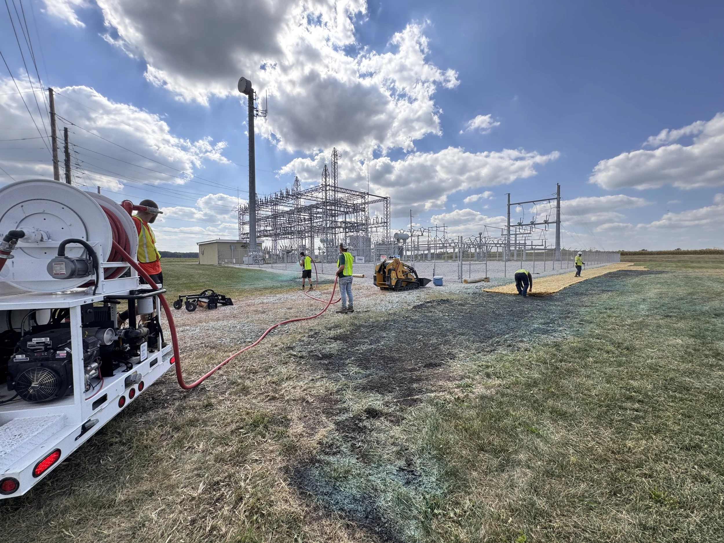 Workers in safety vests and helmets laying gravel near an electrical substation under a partly cloudy sky.