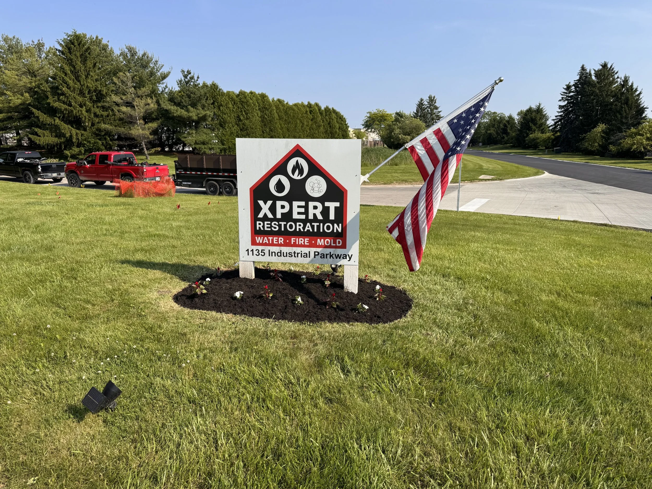 Sign for XPERT Restoration with water, fire, and mold services, located at 1135 Industrial Parkway, beside an American flag on a grassy area near a sidewalk and road, with trucks parked in the background and trees behind.